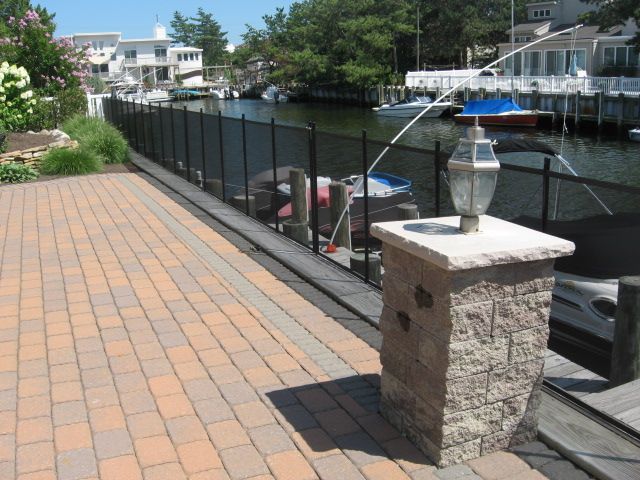 Brick walkway beside canal with black fence, boats, and waterfront houses on a sunny day