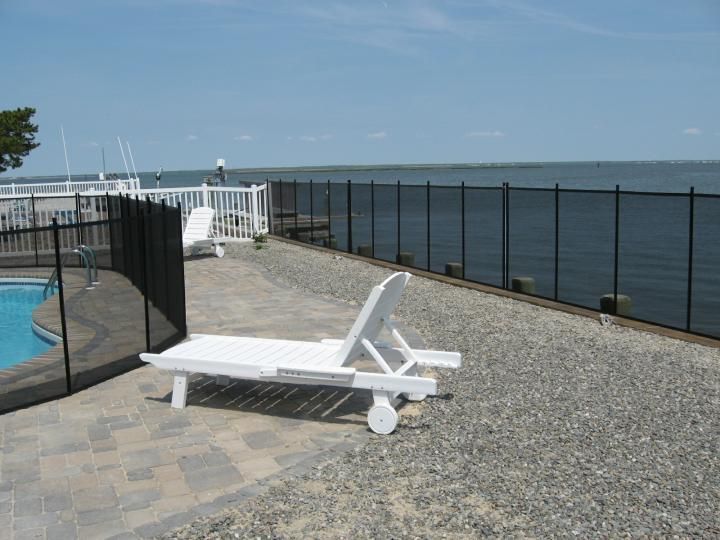 White lounge chair on a gravel patio by a fenced waterfront pool under a clear sky