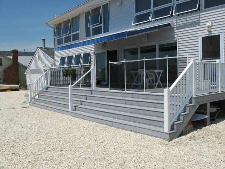 White seaside house with a large gray deck, railings, and sliding glass doors on a gravel lot