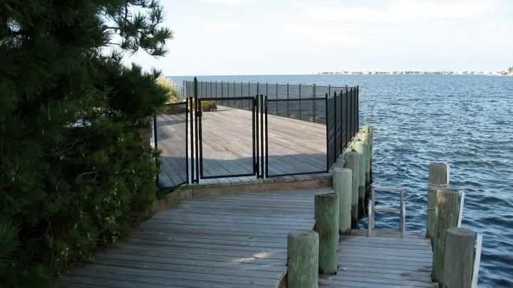 Wooden dock leading to a gated walkway beside a calm body of water under a bright sky