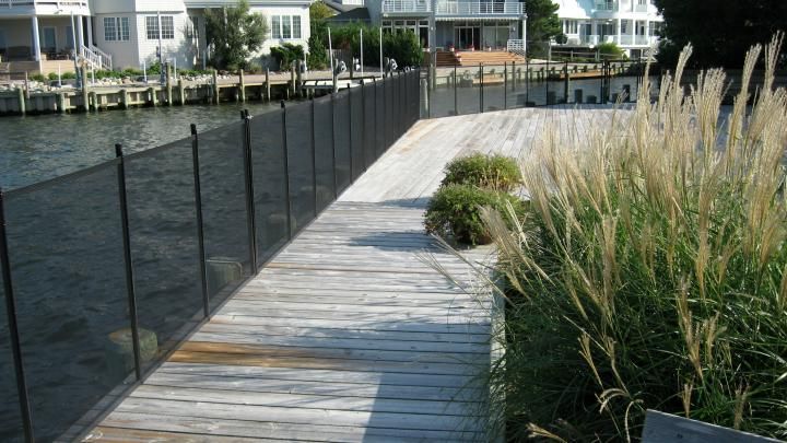 Wooden boardwalk beside dark water, with tall grasses and waterfront homes in the distance.