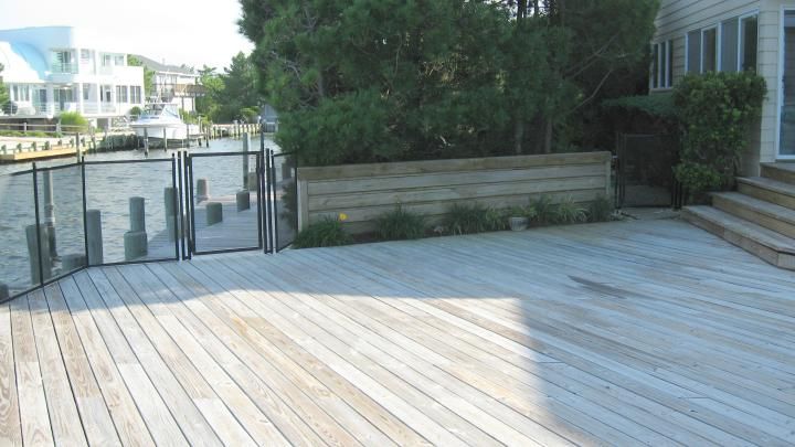 Wooden deck beside a waterfront gate and shrubs, with buildings in the background.