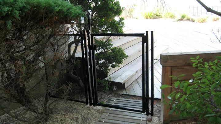 Black metal gate at the top of a narrow stone stairway beside dense green bushes and a wooden deck