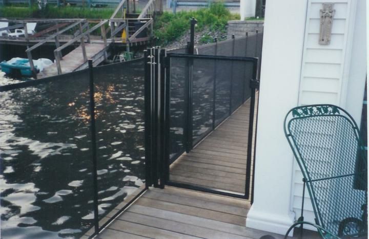 Wooden dock beside black water fencing, with a gate and white house wall on the right.