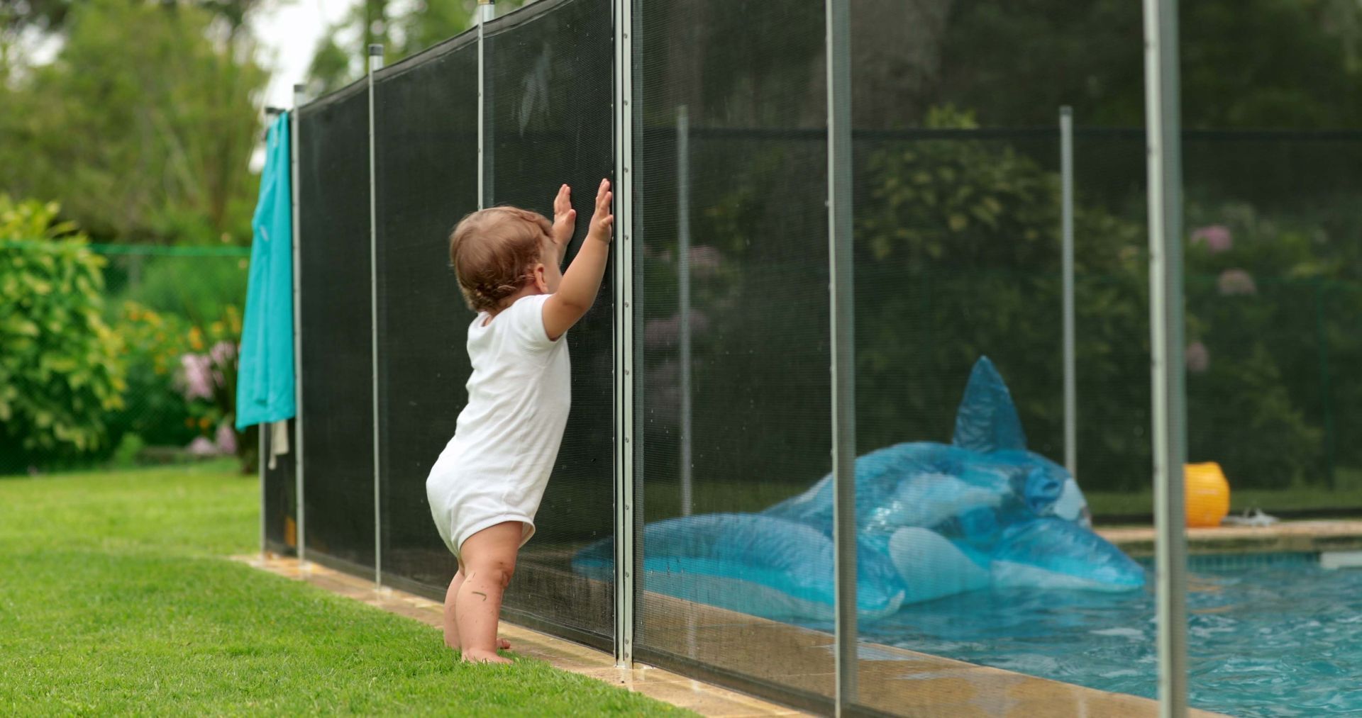 Toddler reaching up at a fence beside a blue tarp in a grassy yard