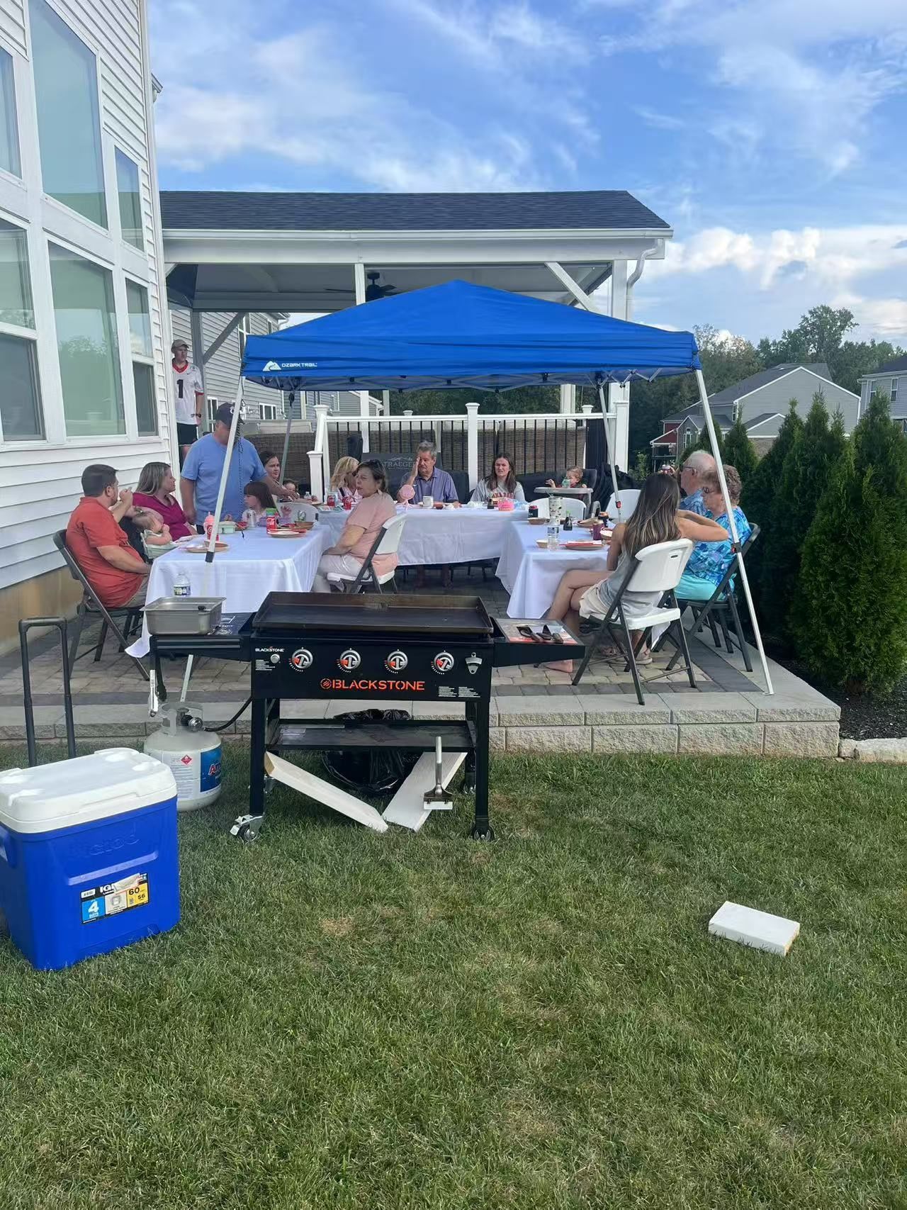 A group of people are sitting at tables under a blue tent in a backyard.