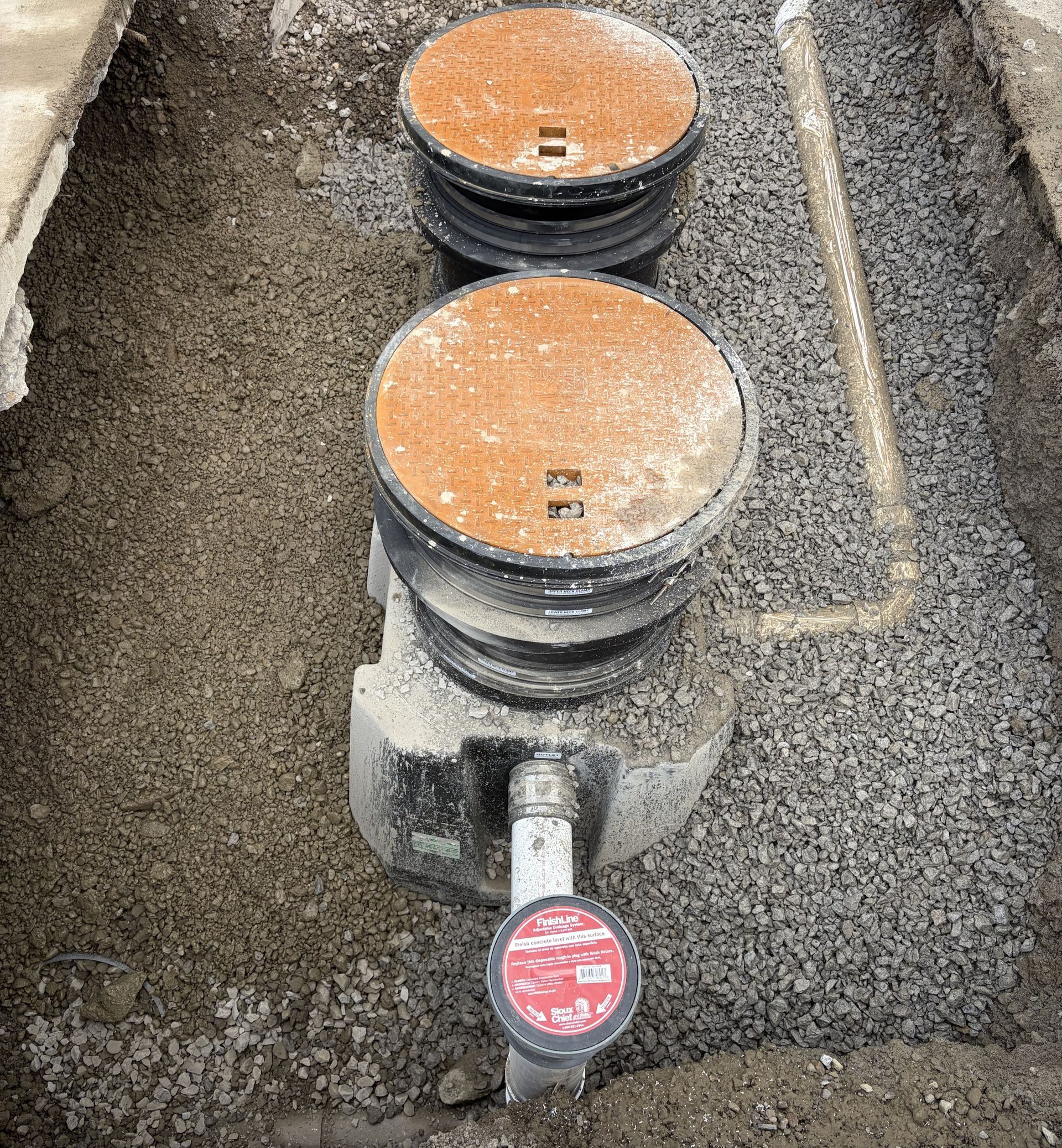 Two round utility meter lids in a gravel trench, one with a red valve cap in front.