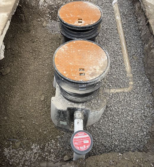 Two round utility meter lids in a gravel trench, one with a red valve cap in front.