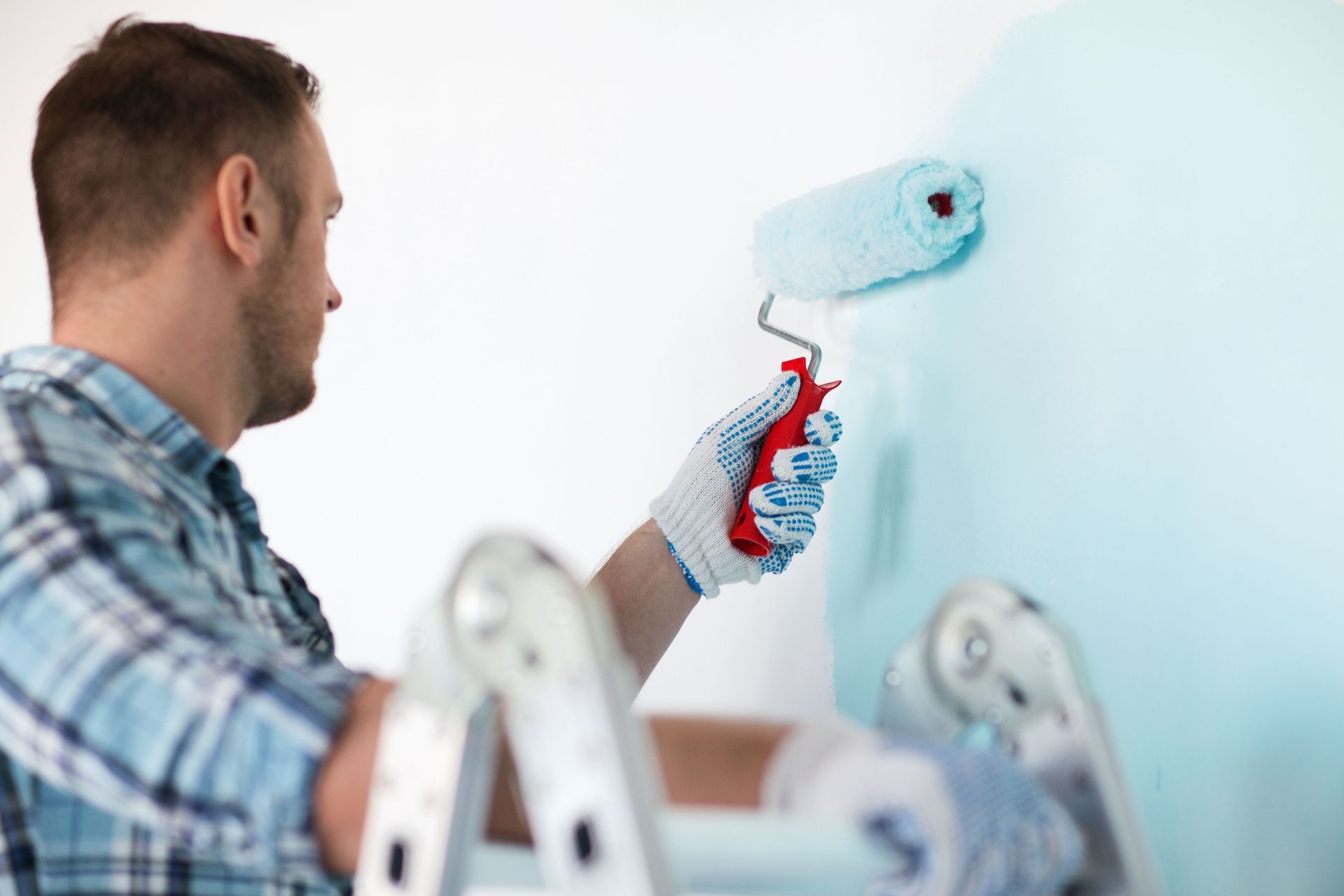 Man in blue plaid shirt painting a wall with a roller; light blue paint.