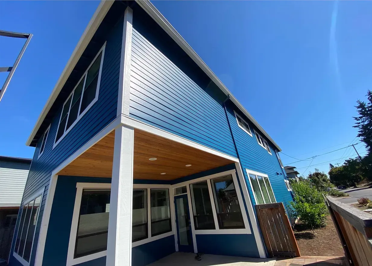 Blue modern house with white trim, large windows, and wooden porch ceiling against a clear blue sky.