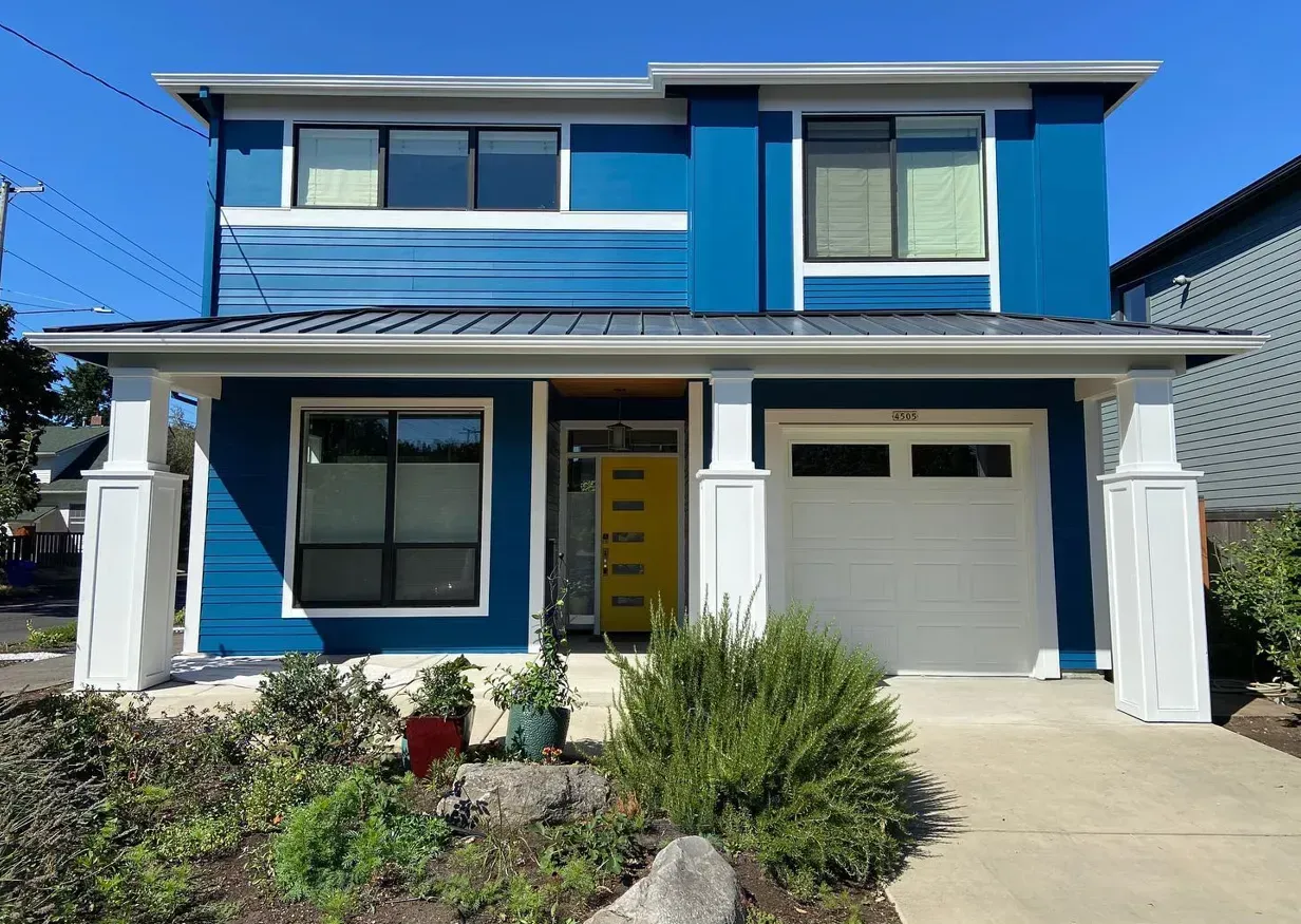 Blue two-story house with white trim, a yellow door, and a white garage door. Landscaping in front.