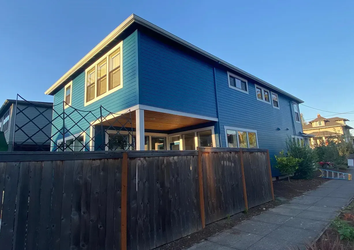 Blue two-story house with covered patio behind a wooden fence, sidewalk, and sunny sky.