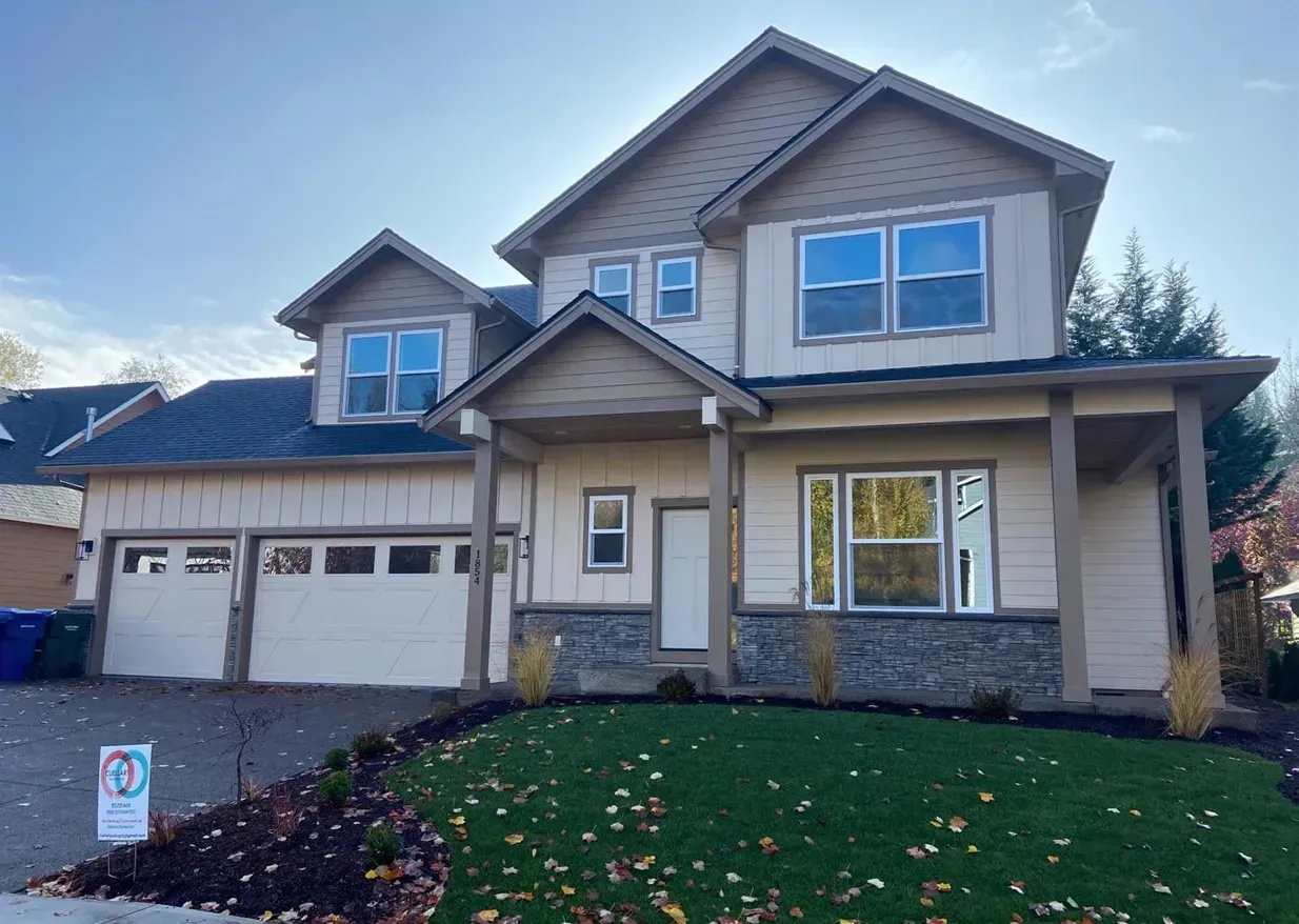 Two-story beige house with a stone facade, garage, porch, and green lawn under a blue sky.