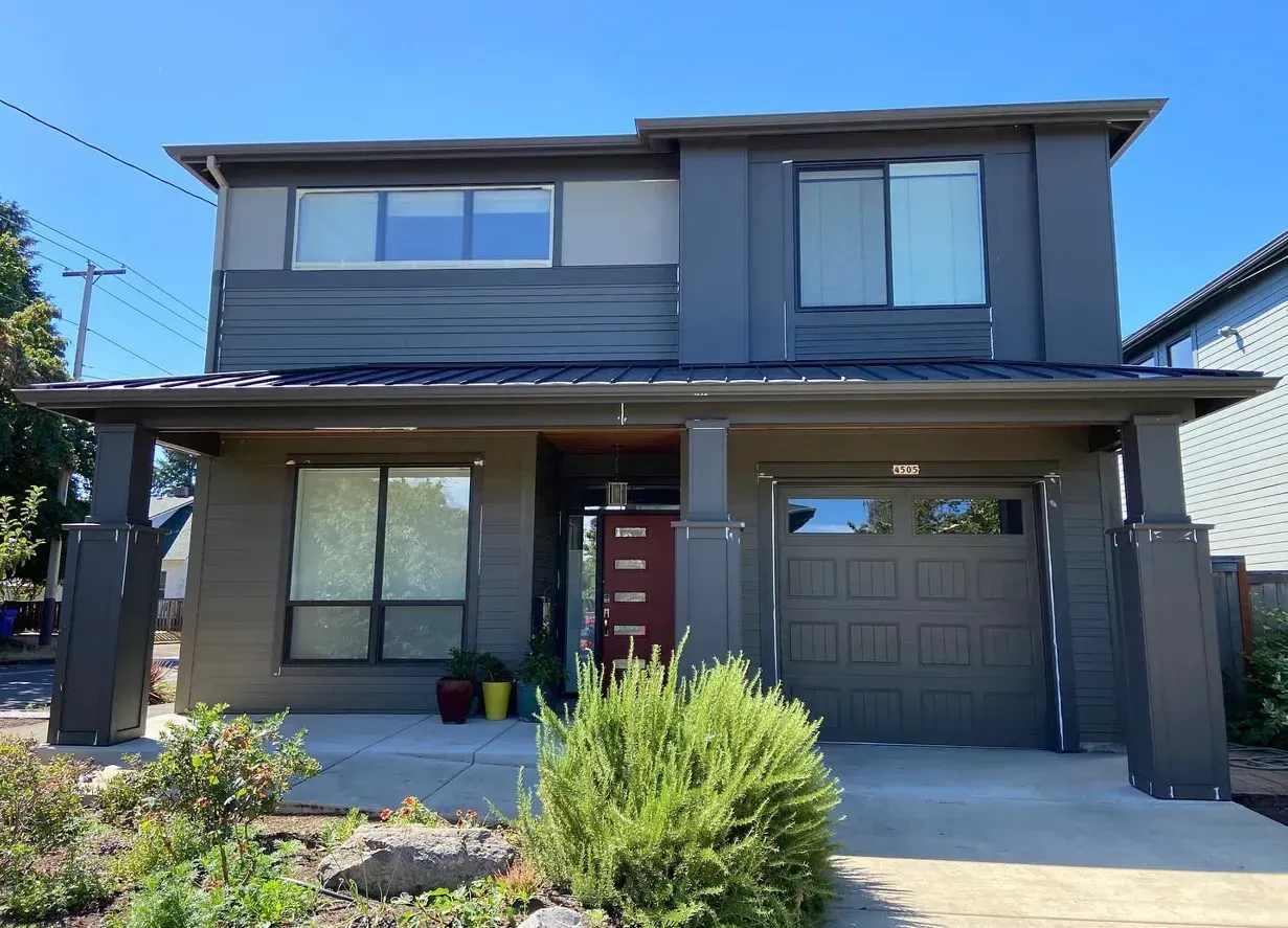 Modern two-story house with gray and brown exterior paint. Garage door and front porch visible. Sunny day.