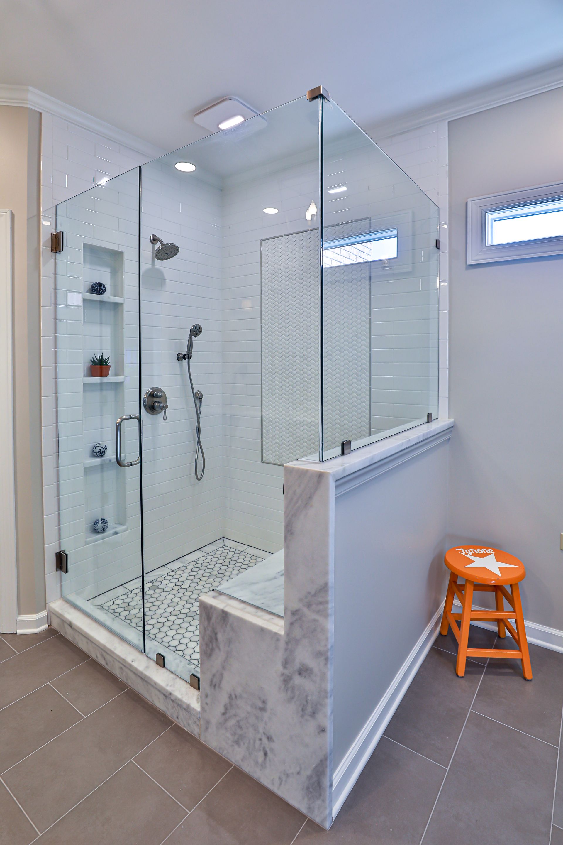 Glass-enclosed walk-in shower with marble accent, gray walls, and an orange stool next to it.
