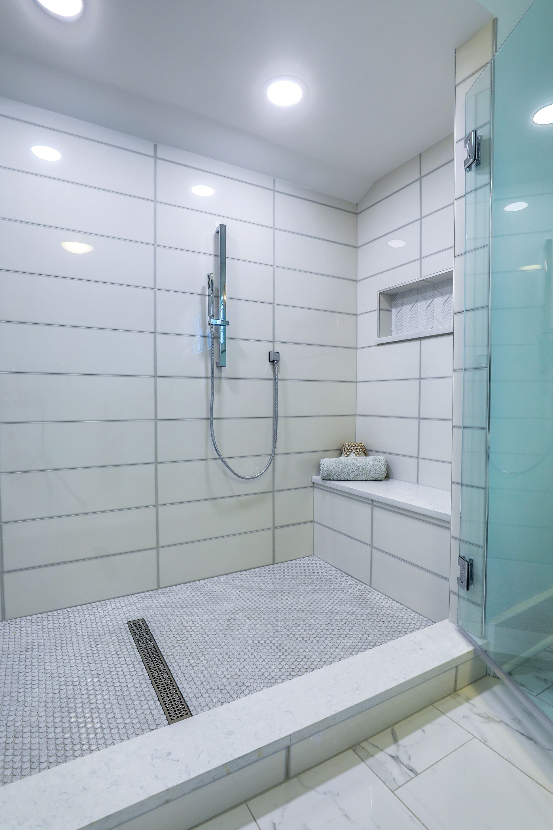 White-tiled shower with a built-in bench, glass door, and pebble-tiled floor.