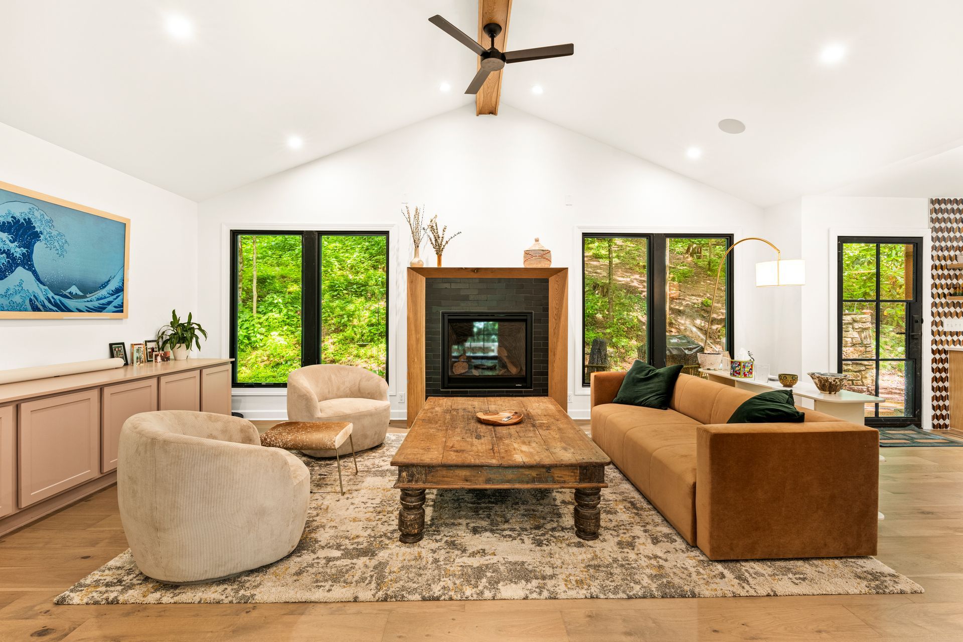 A modern living room with a fireplace, brown sofa, beige chairs, and large windows overlooking trees.
