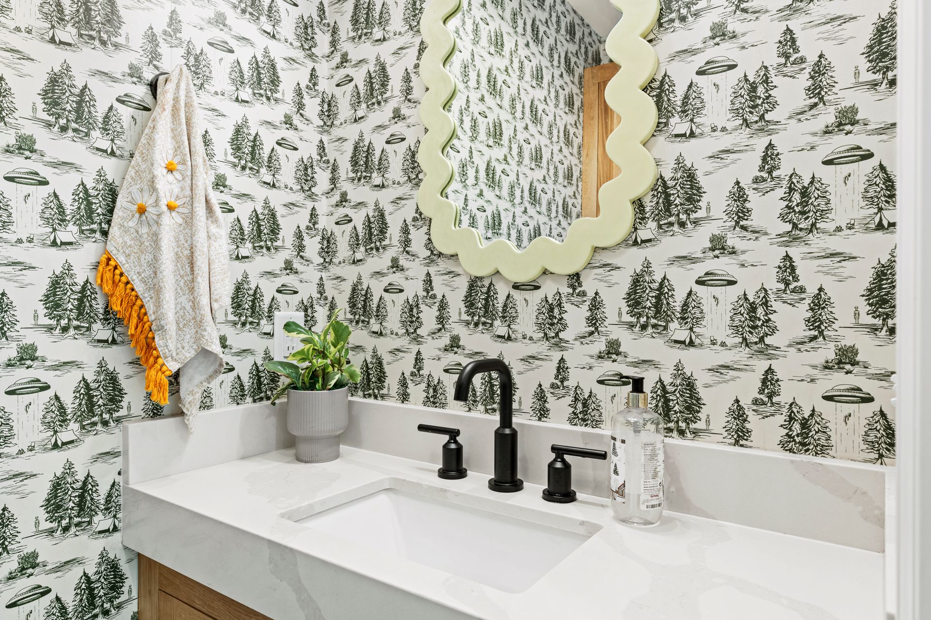 Bathroom with forest-patterned wallpaper, white countertop, black faucet, scalloped mirror, and hand towel.