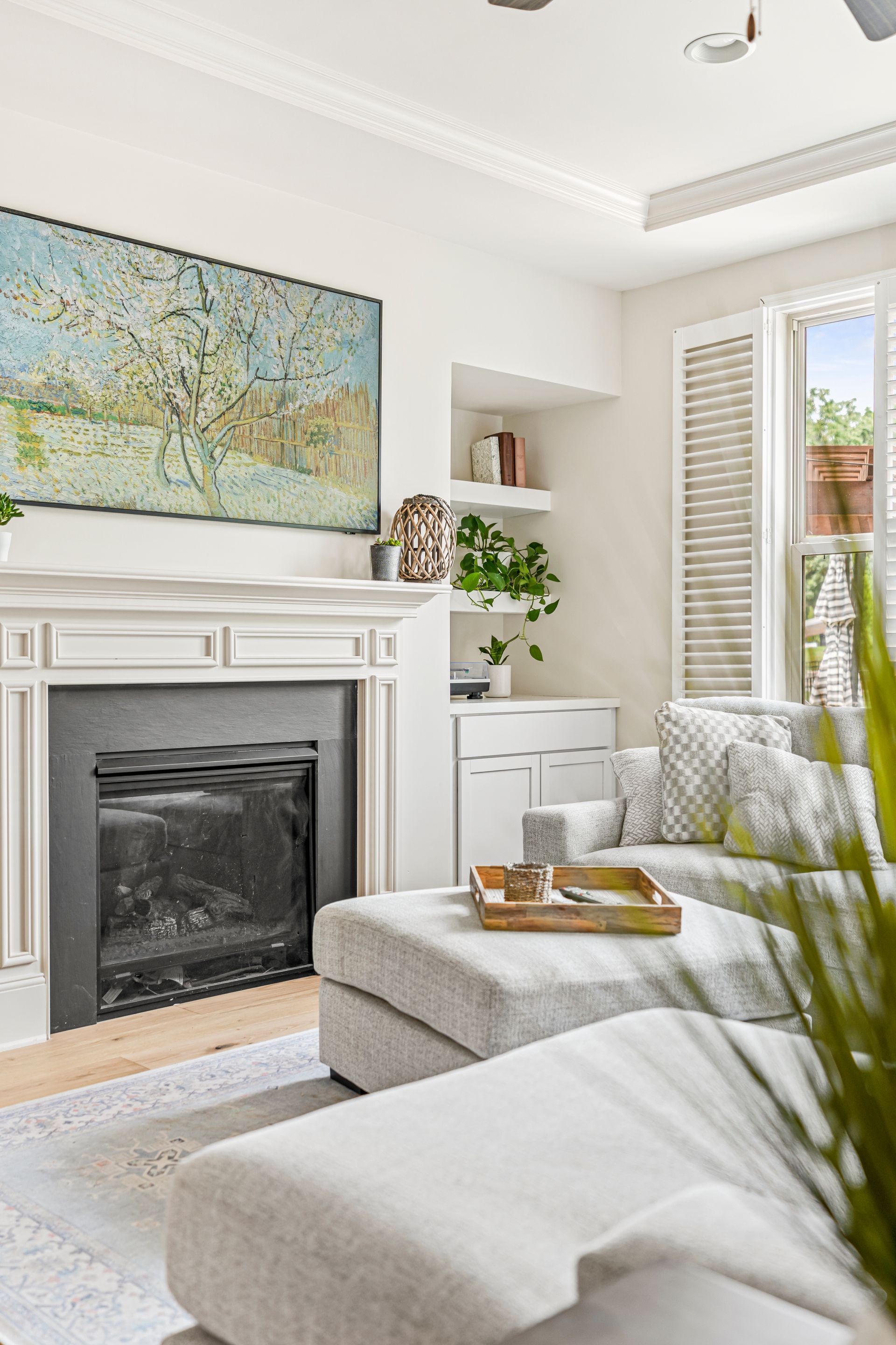 Cozy living room with fireplace, gray couch, and artwork. Sunlight streams through a shuttered window.