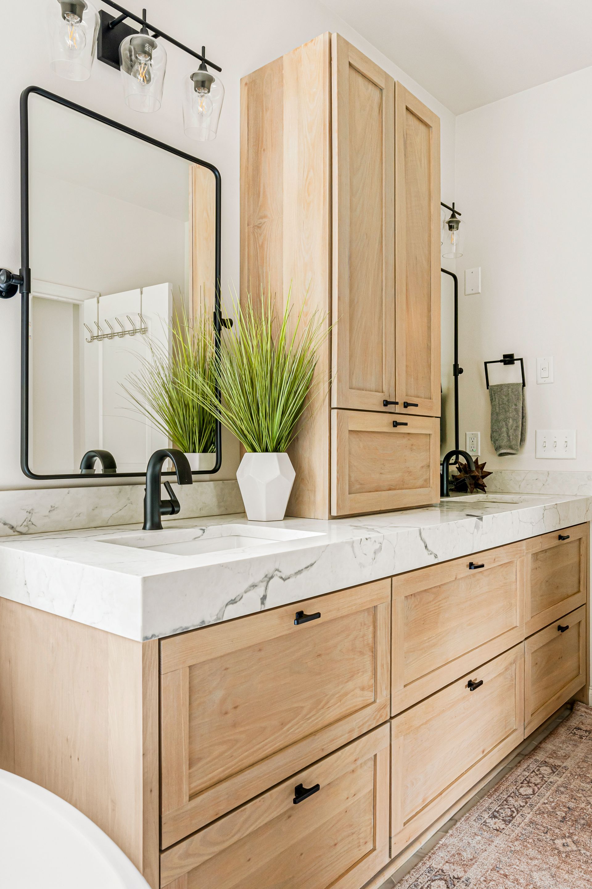 Bathroom with light wood cabinets, marble countertop, black fixtures, and a tall storage unit.