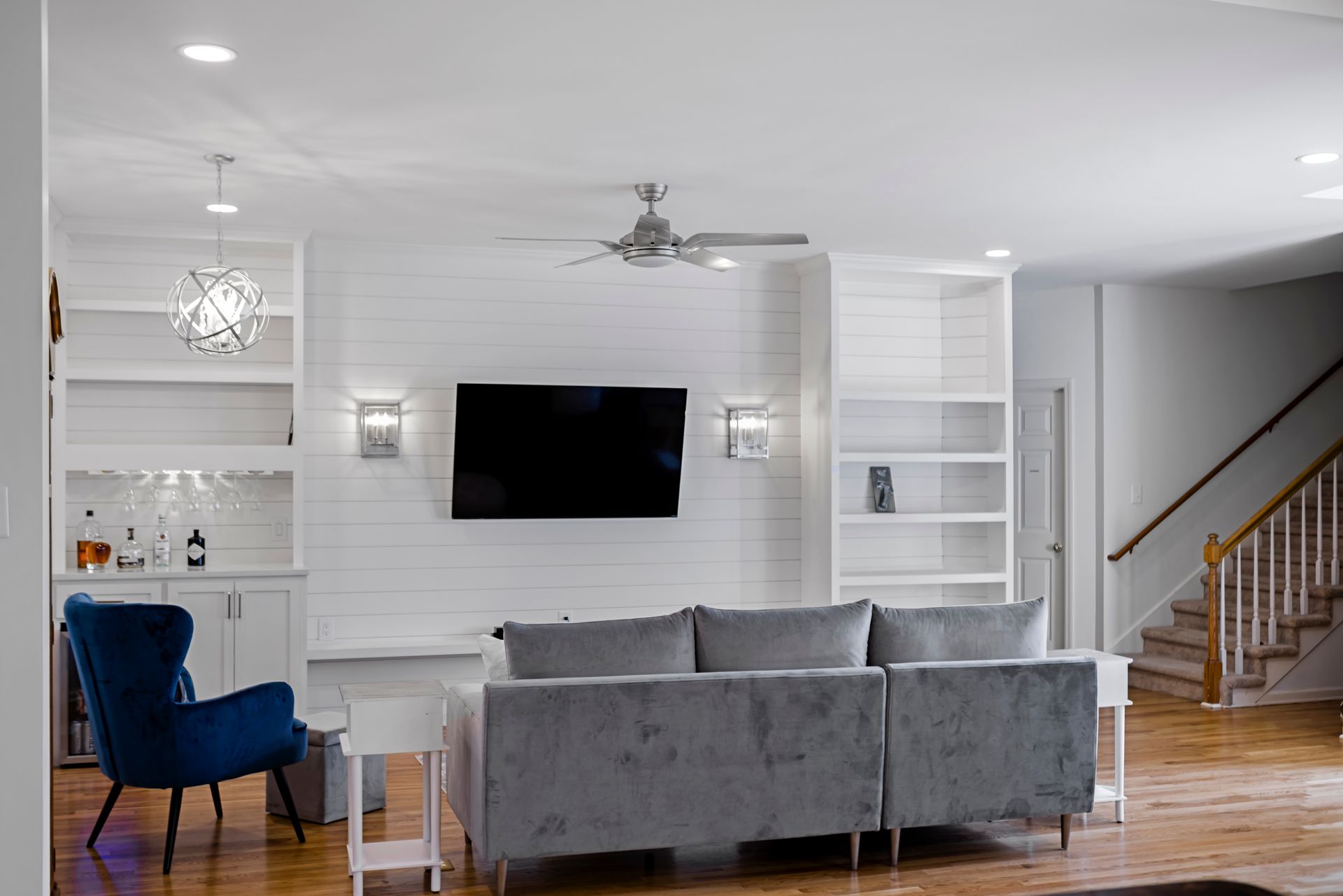 Living room with white shiplap wall, mounted TV, gray sectional, blue chair, and built-in shelving.