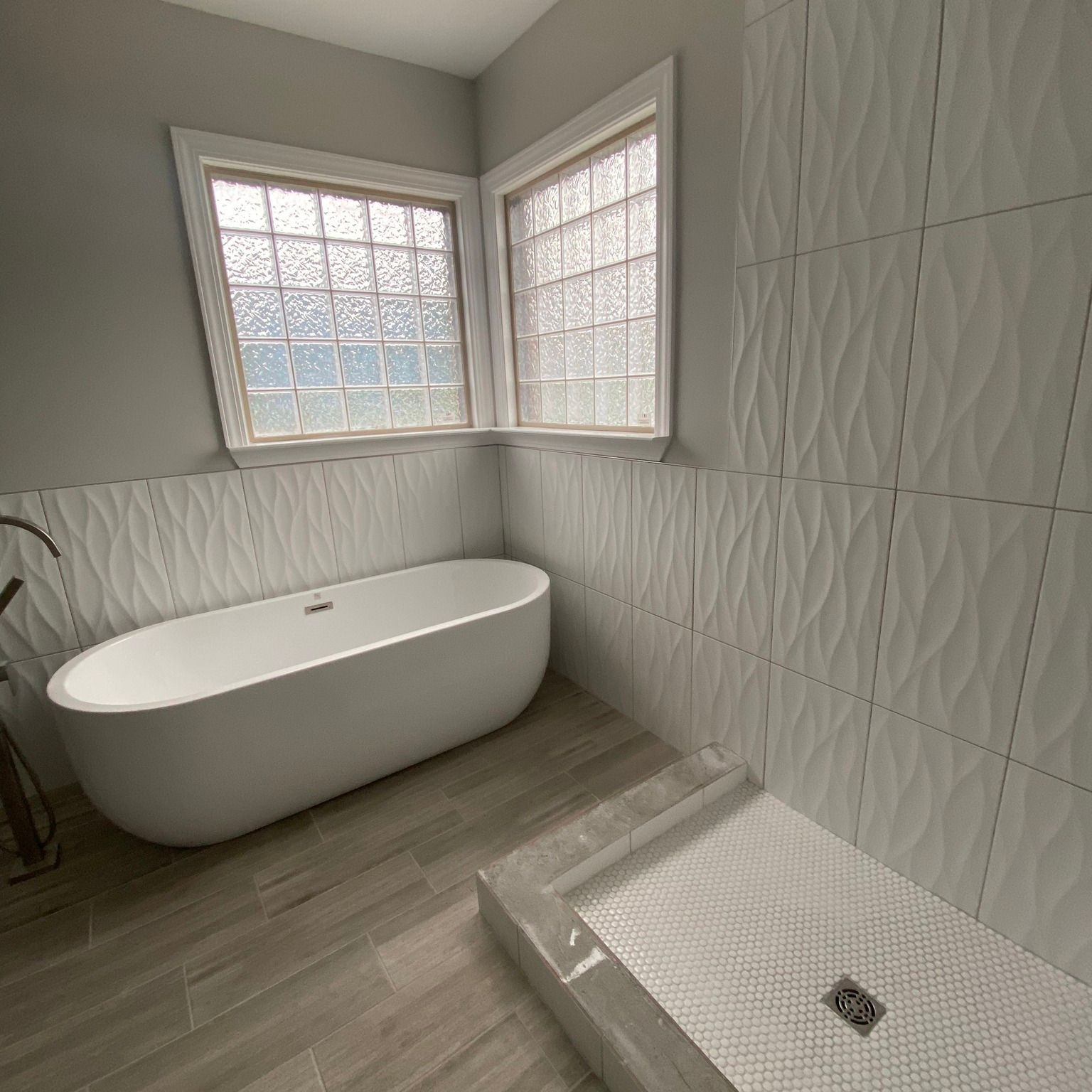 Modern bathroom with a white tub, shower, and textured white tile walls. Wooden floor.
