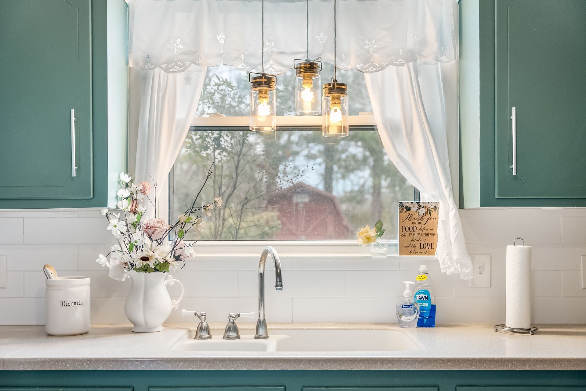 Kitchen sink with flowers, window with a view of a red barn, and teal cabinets.