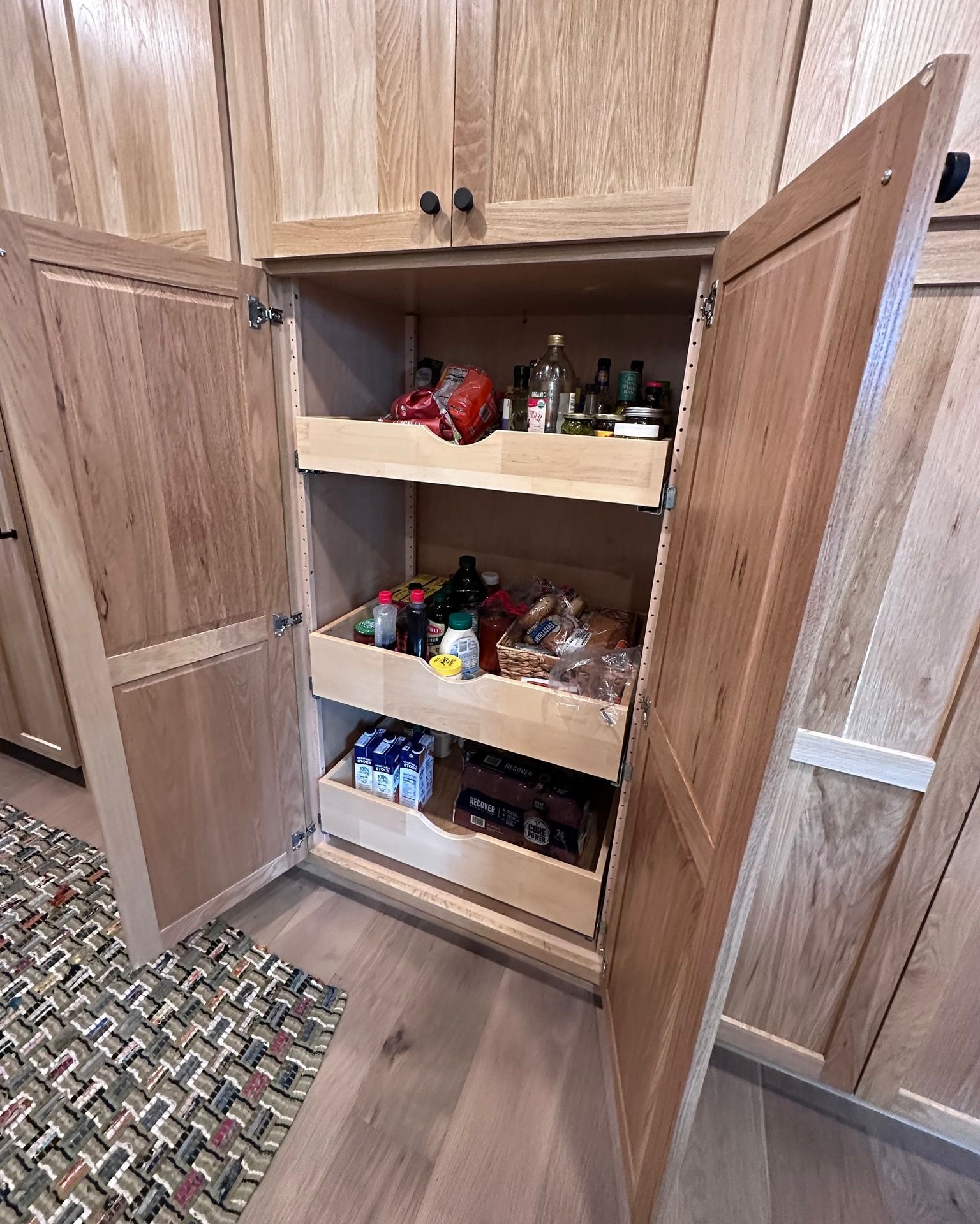 Wooden pantry cabinet with open doors, showing pull-out shelves filled with food items.