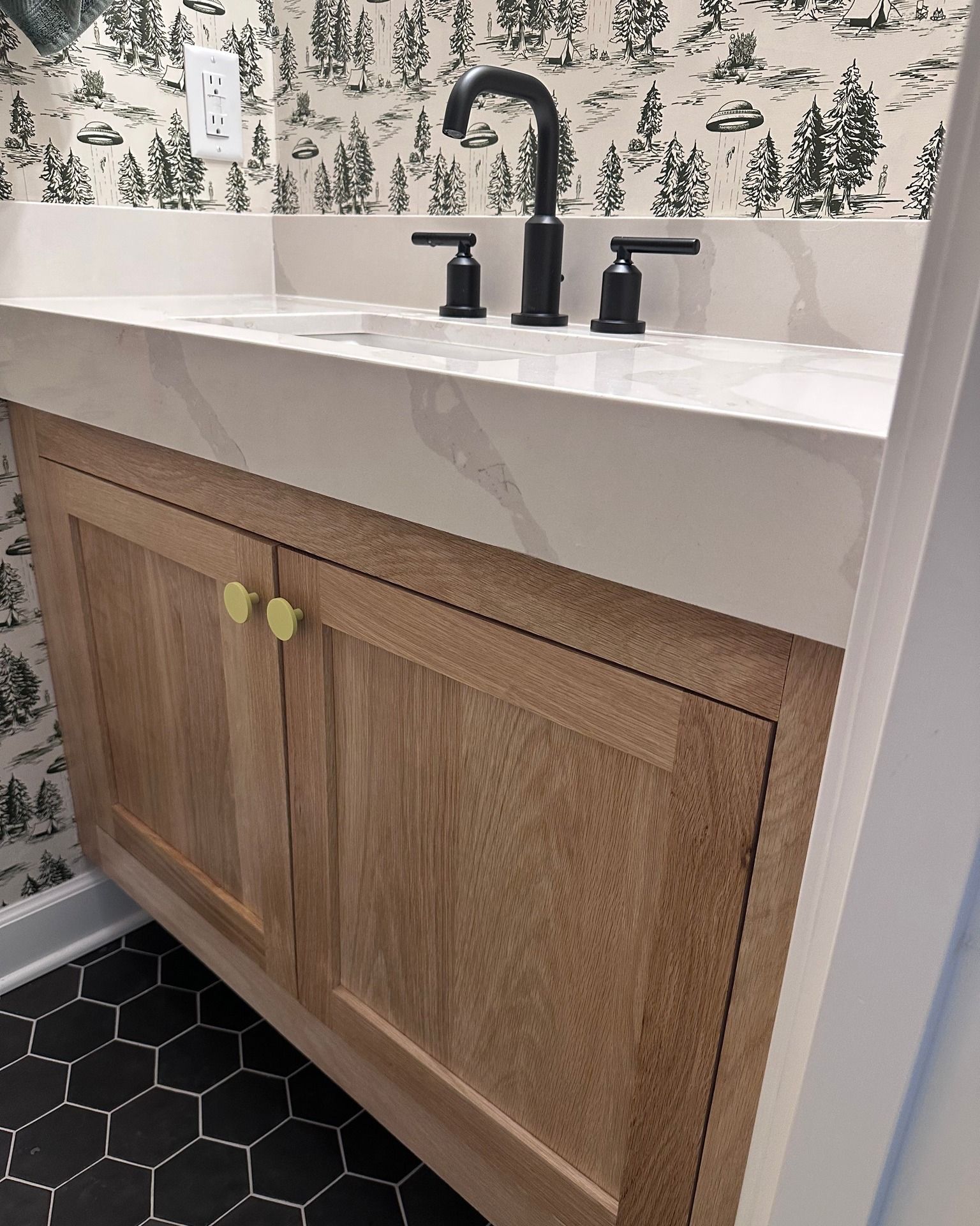 Bathroom vanity with light wood cabinets, white countertop, black faucet, gold knobs, and black and white wallpaper.
