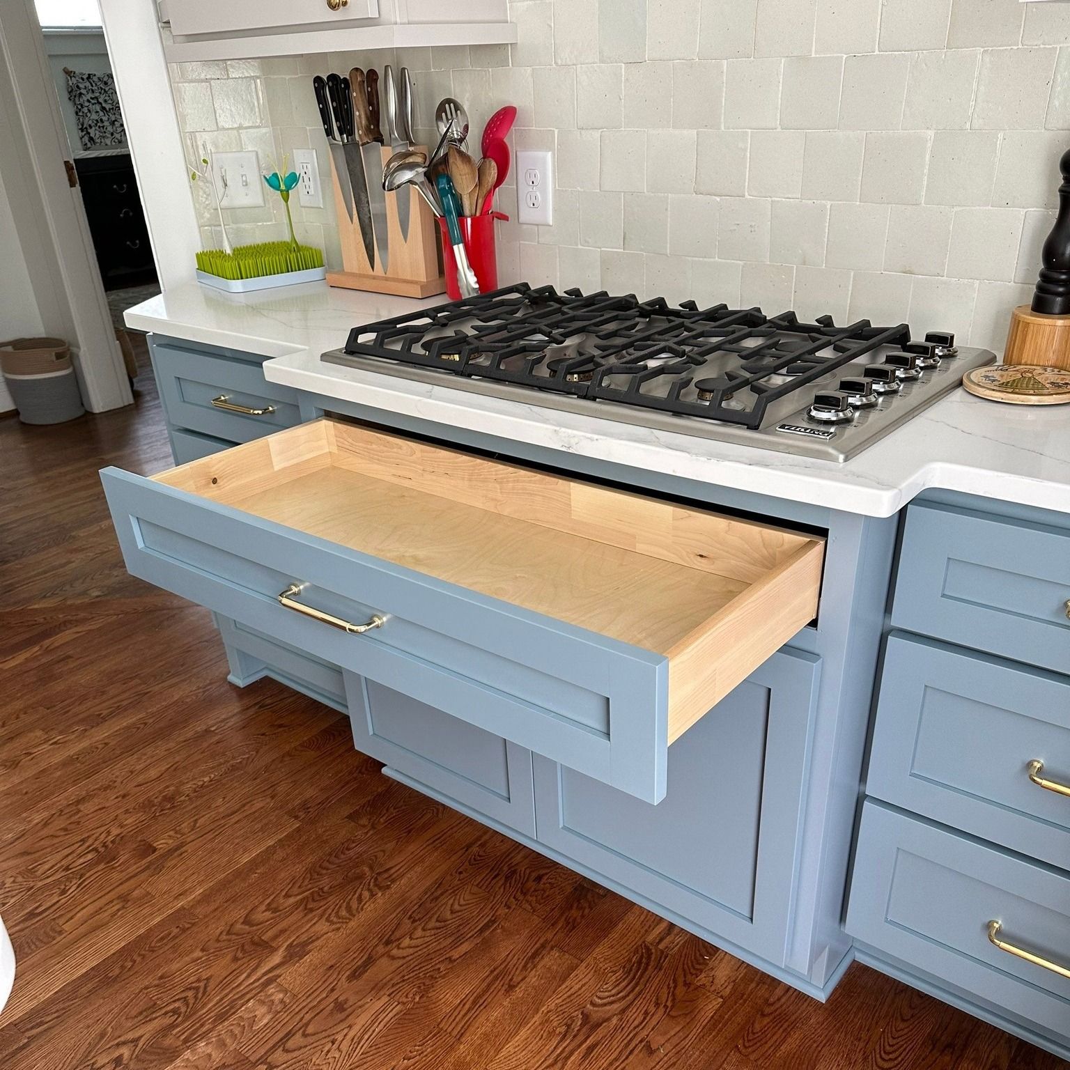 Open blue kitchen drawer under a gas cooktop; wooden floor, gray cabinets, gold hardware.