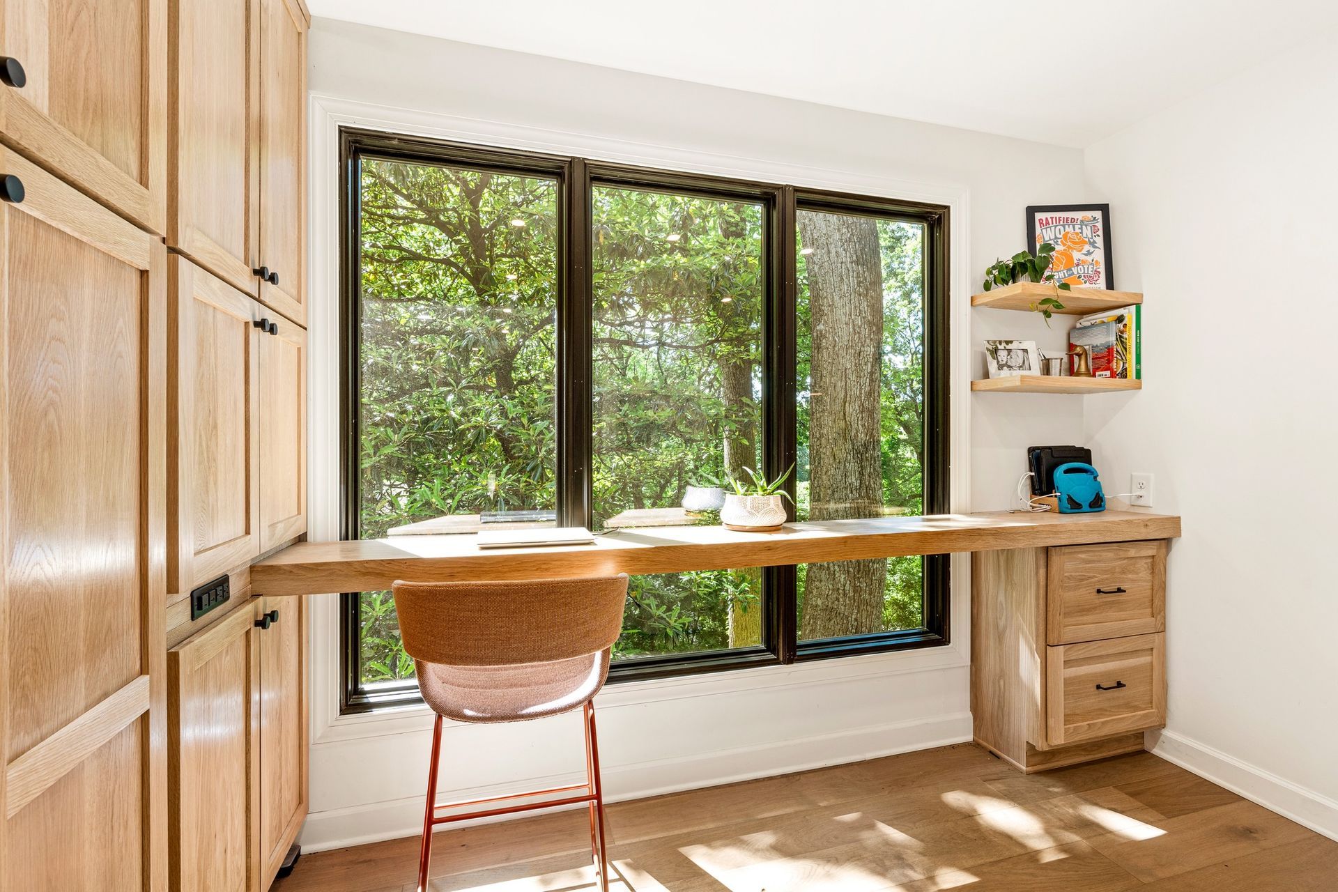 Desk area with large window overlooking trees, wooden desk, cabinets, and a chair.