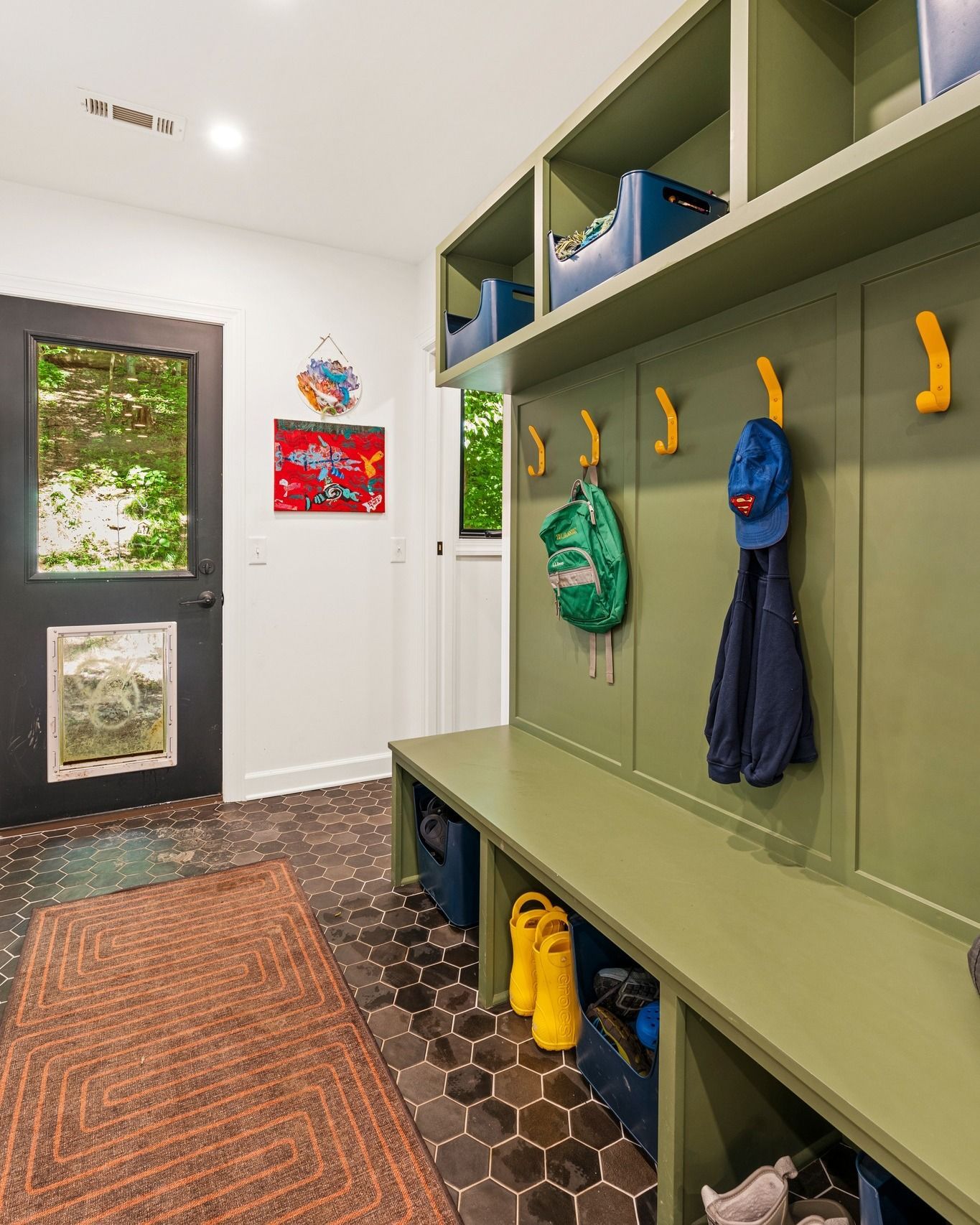 Green-painted entryway with built-in storage, bench, and hooks. A blue hat, coat, and backpack hang.