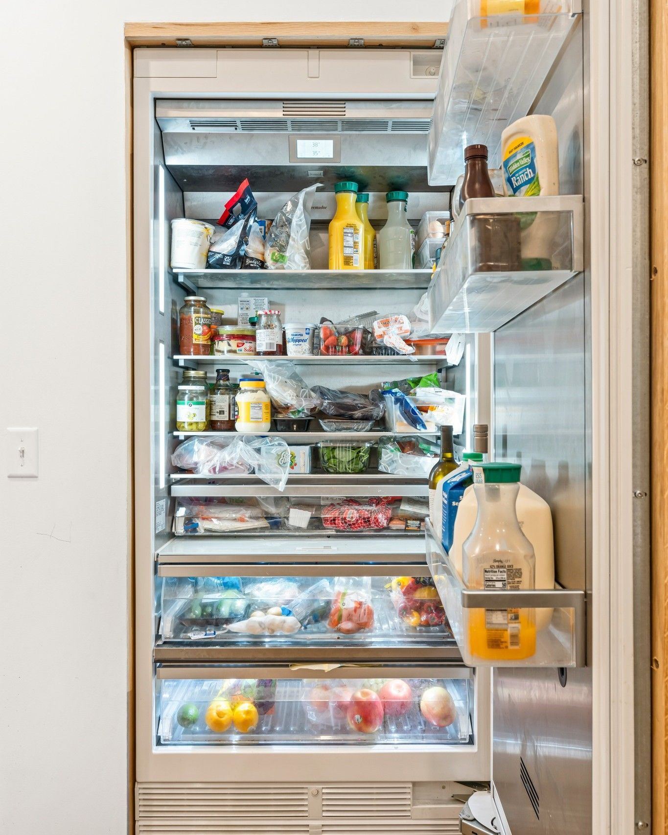 Refrigerator open, packed with food.  Produce, condiments, and containers fill the shelves and door storage.
