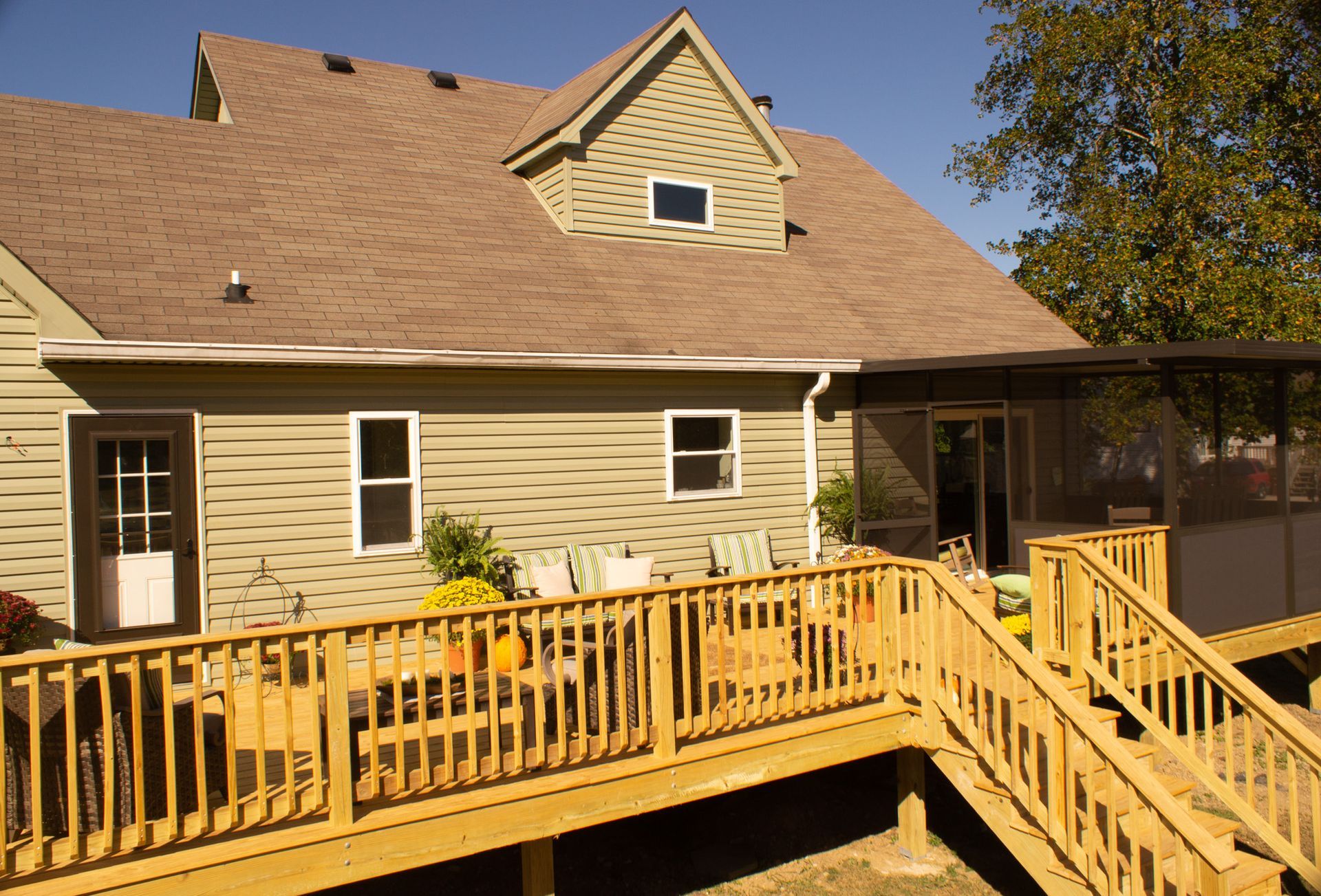 Wooden deck with railing and steps leading to a house with a screened porch; green siding, brown roof, sunny day.