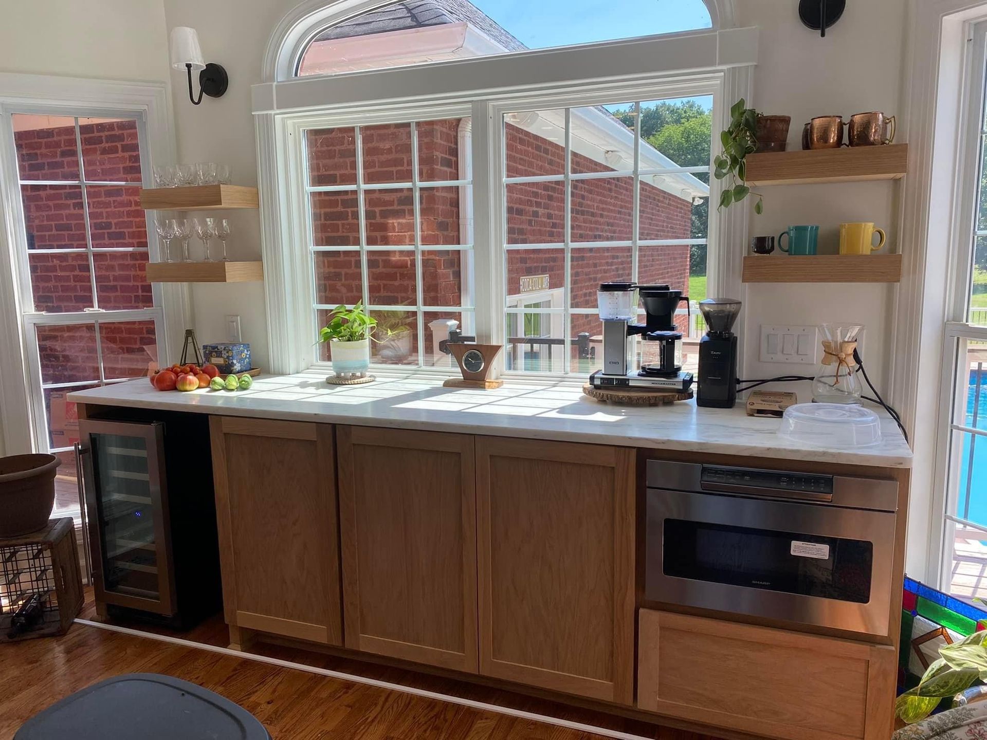 A light-filled kitchen with a brick exterior view, featuring cabinets, a wine cooler, coffee maker, and shelves.
