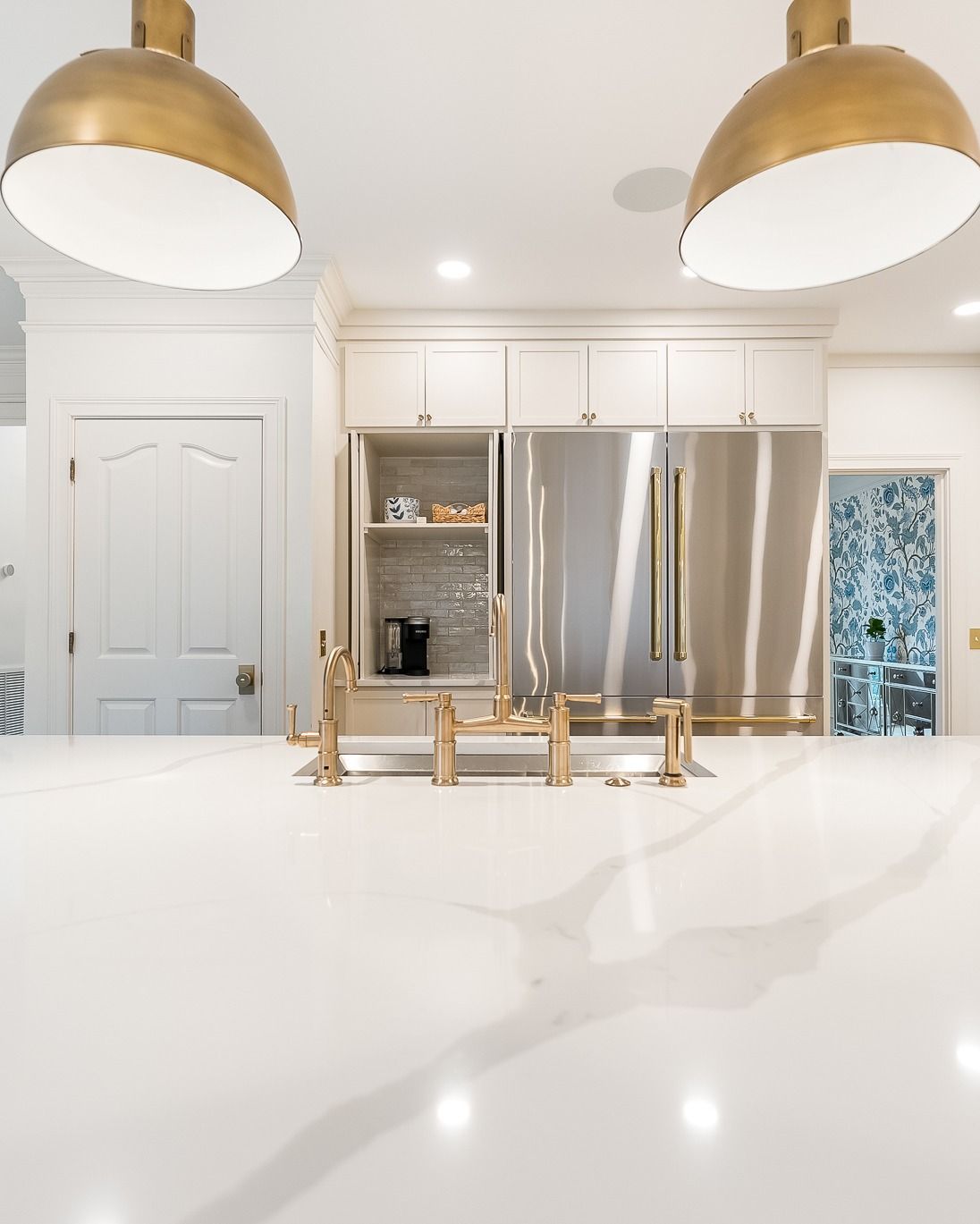 Bright kitchen with white countertop, stainless steel fridge, and gold light fixtures.