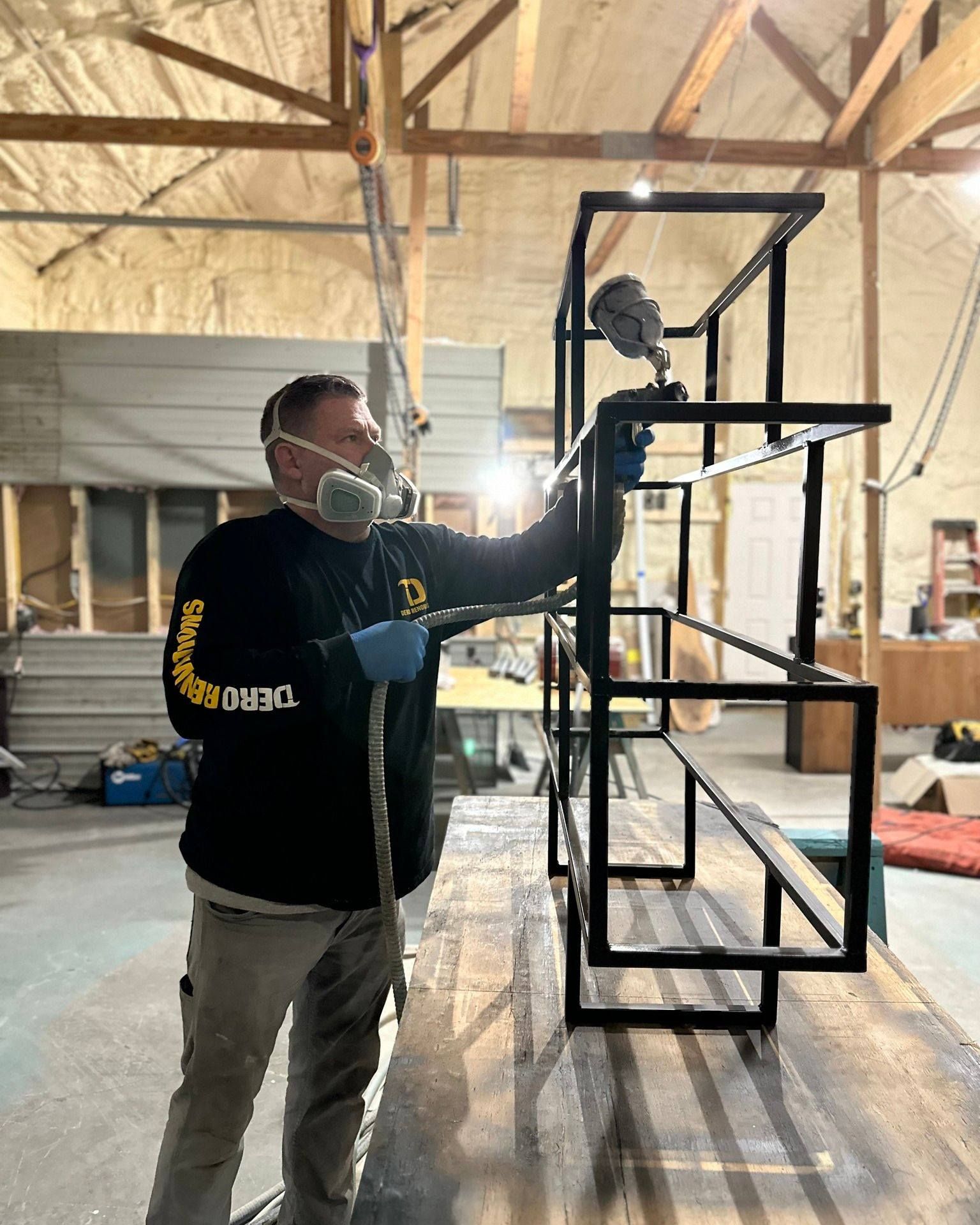 Man sprays black paint on metal shelving unit inside a workshop.