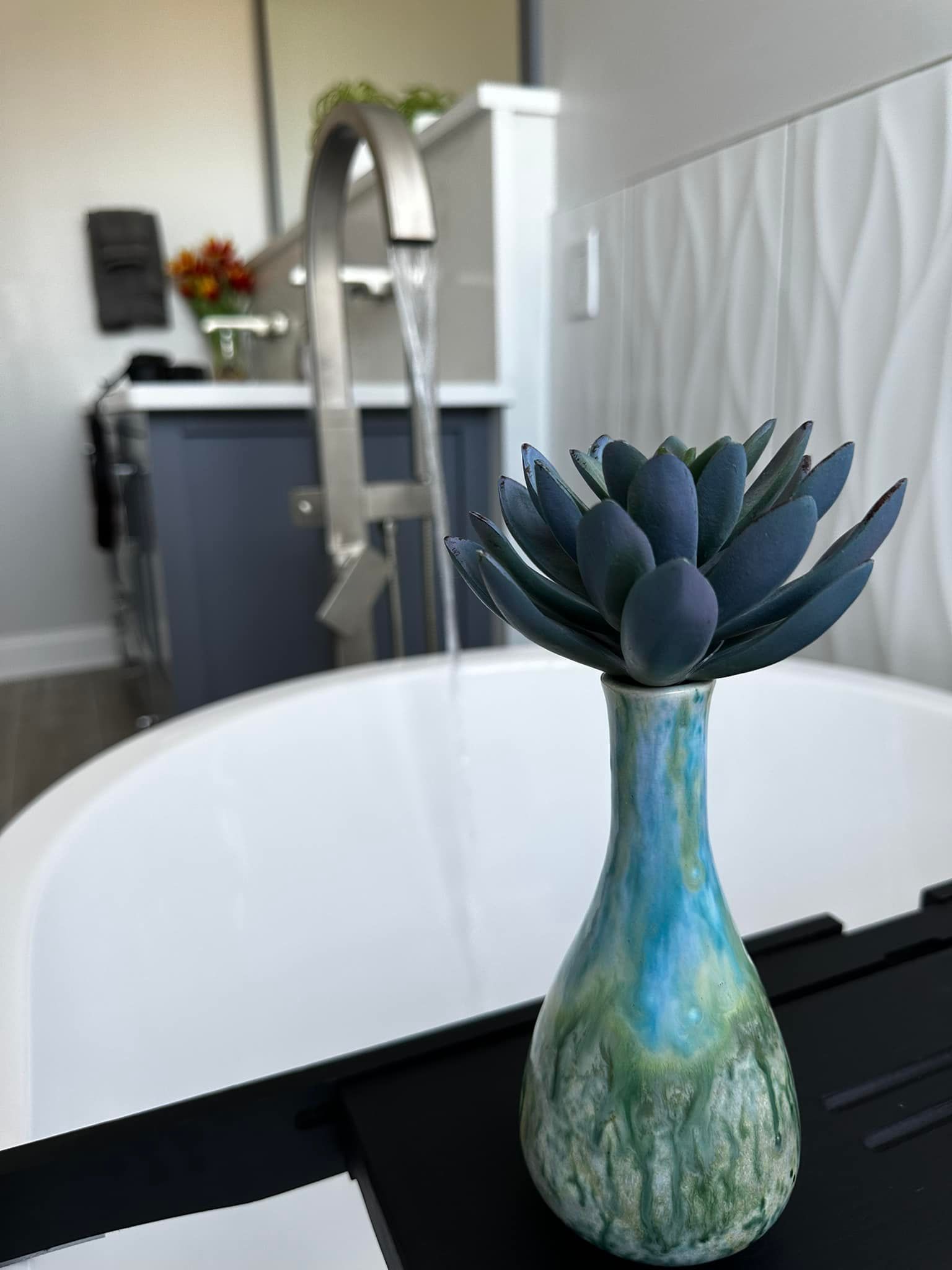 A blue faux succulent in a colorful vase sits on a black tray over a white bathtub with running water.
