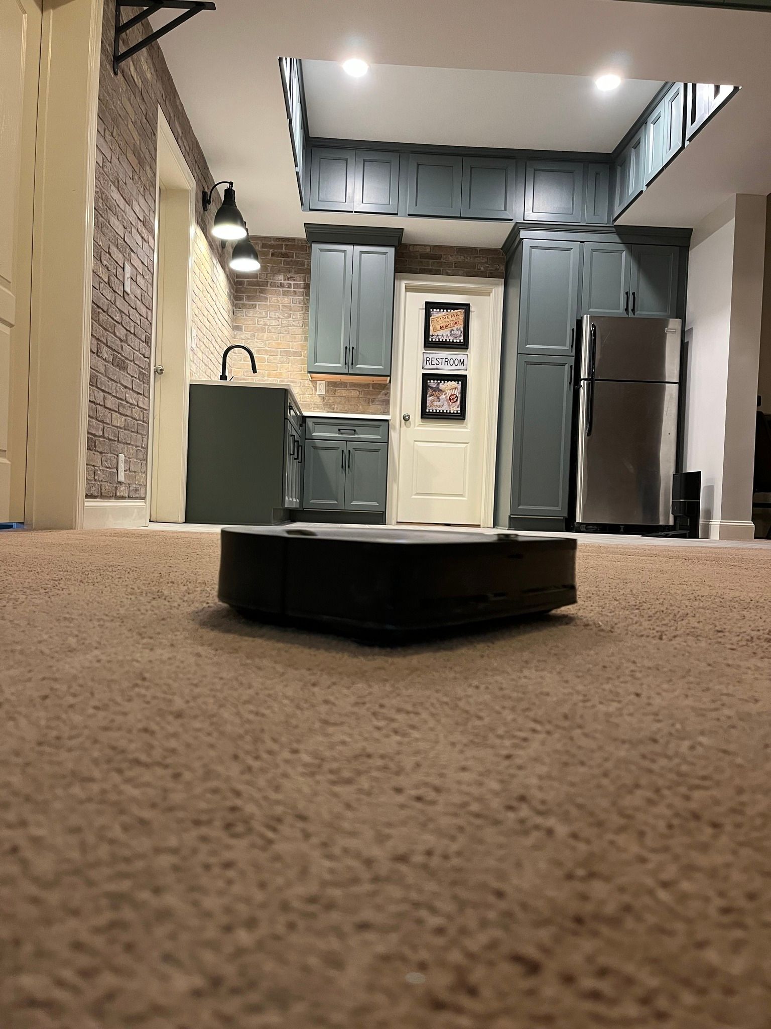 Robot vacuum on brown carpet; kitchen in background with gray cabinets, refrigerator, and doorway.