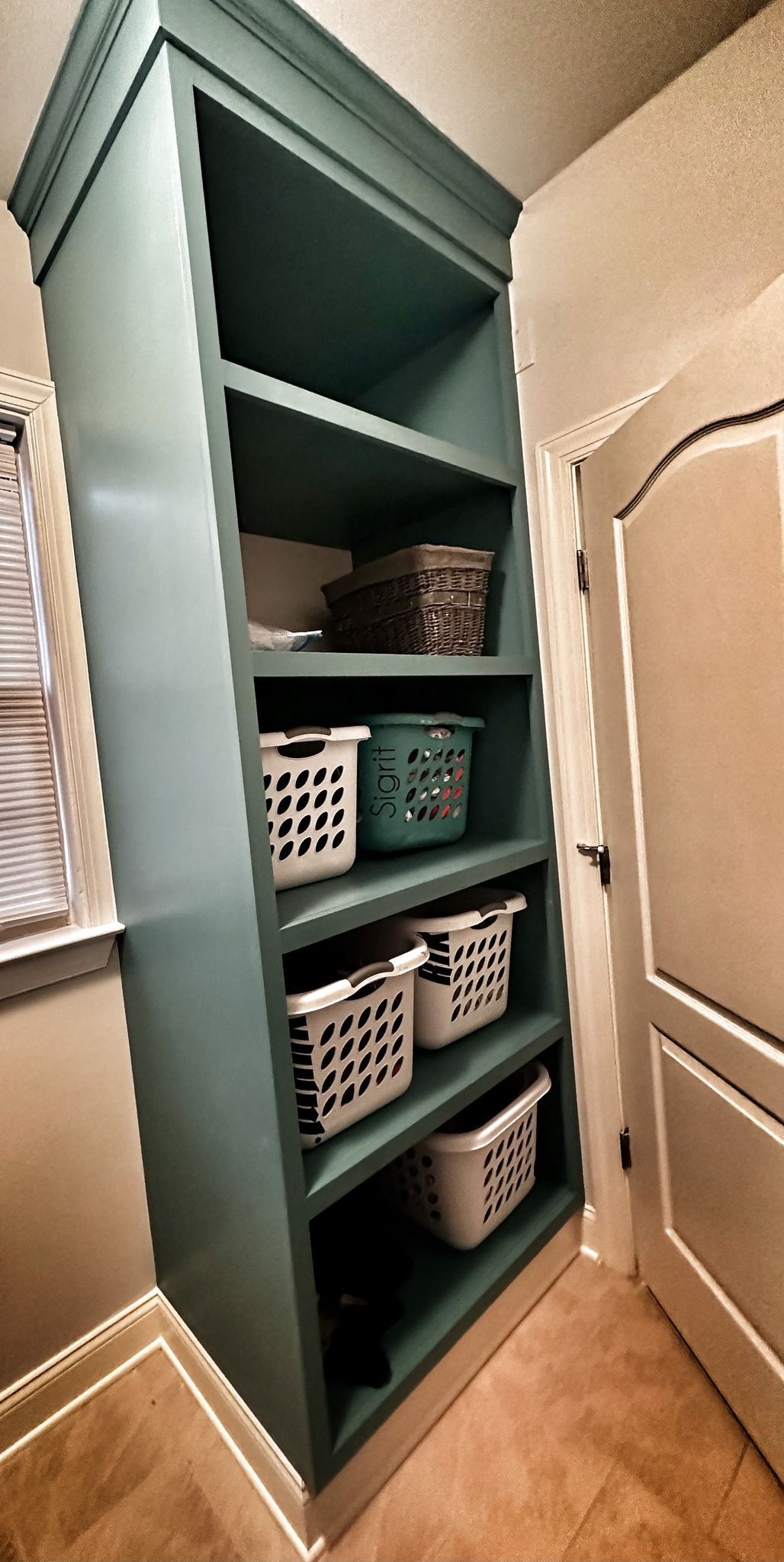 Tall teal shelving unit with laundry baskets, baskets, and door in hallway.