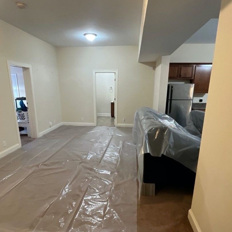 A room with light-colored walls, a clear plastic sheet on the floor, and a couch covered in plastic.