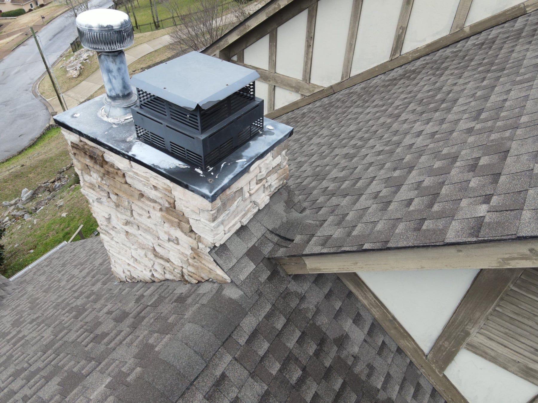 Stone chimney on a shingled roof, topped with a metal cap and vent, in an outdoor setting.