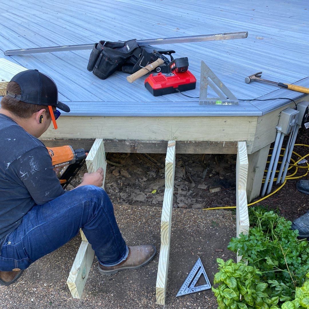 A person builds a deck, using a nail gun on lumber. Tools, including a level, sit nearby.