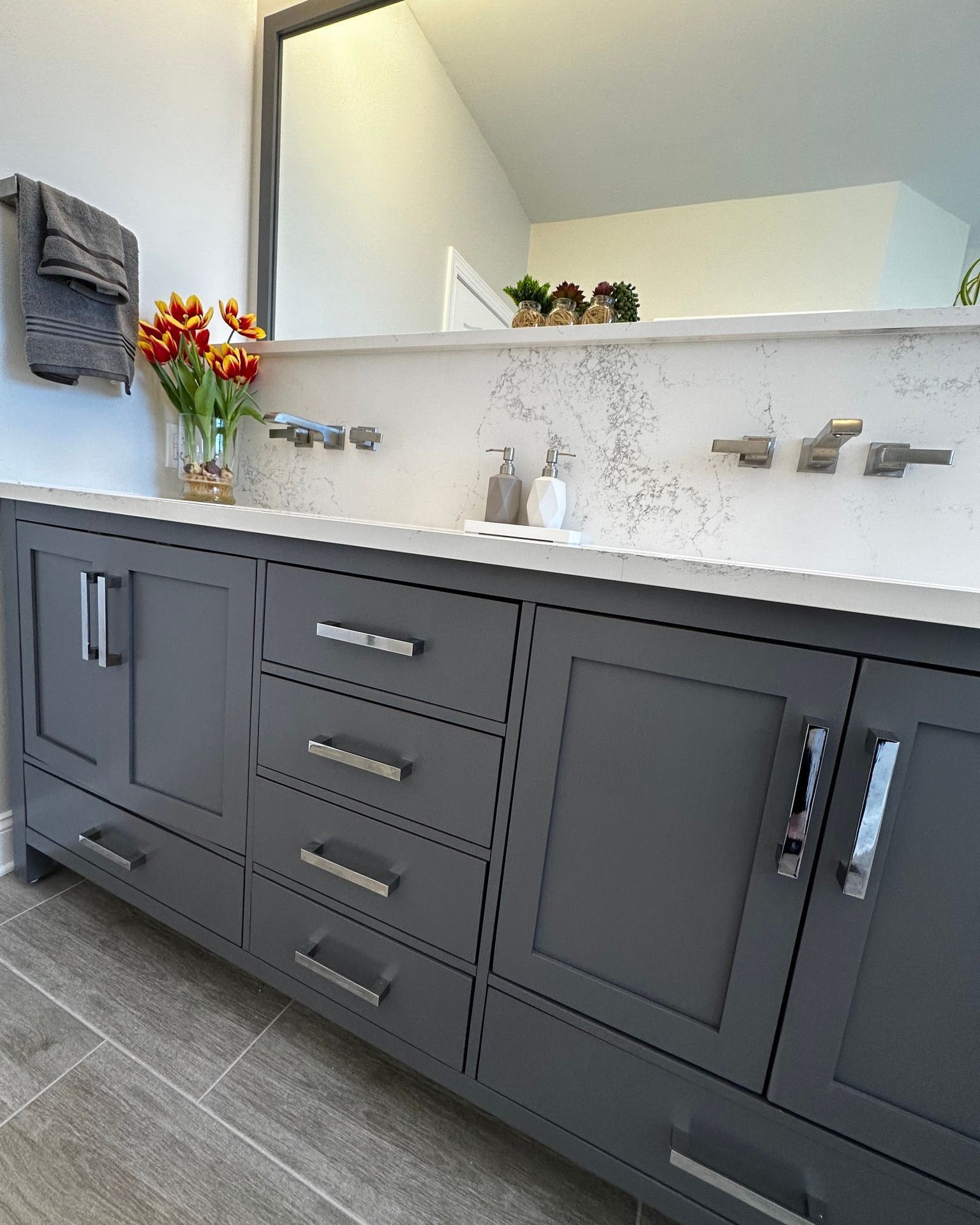 Gray bathroom vanity with silver fixtures, white countertop, and flowers in a vase.