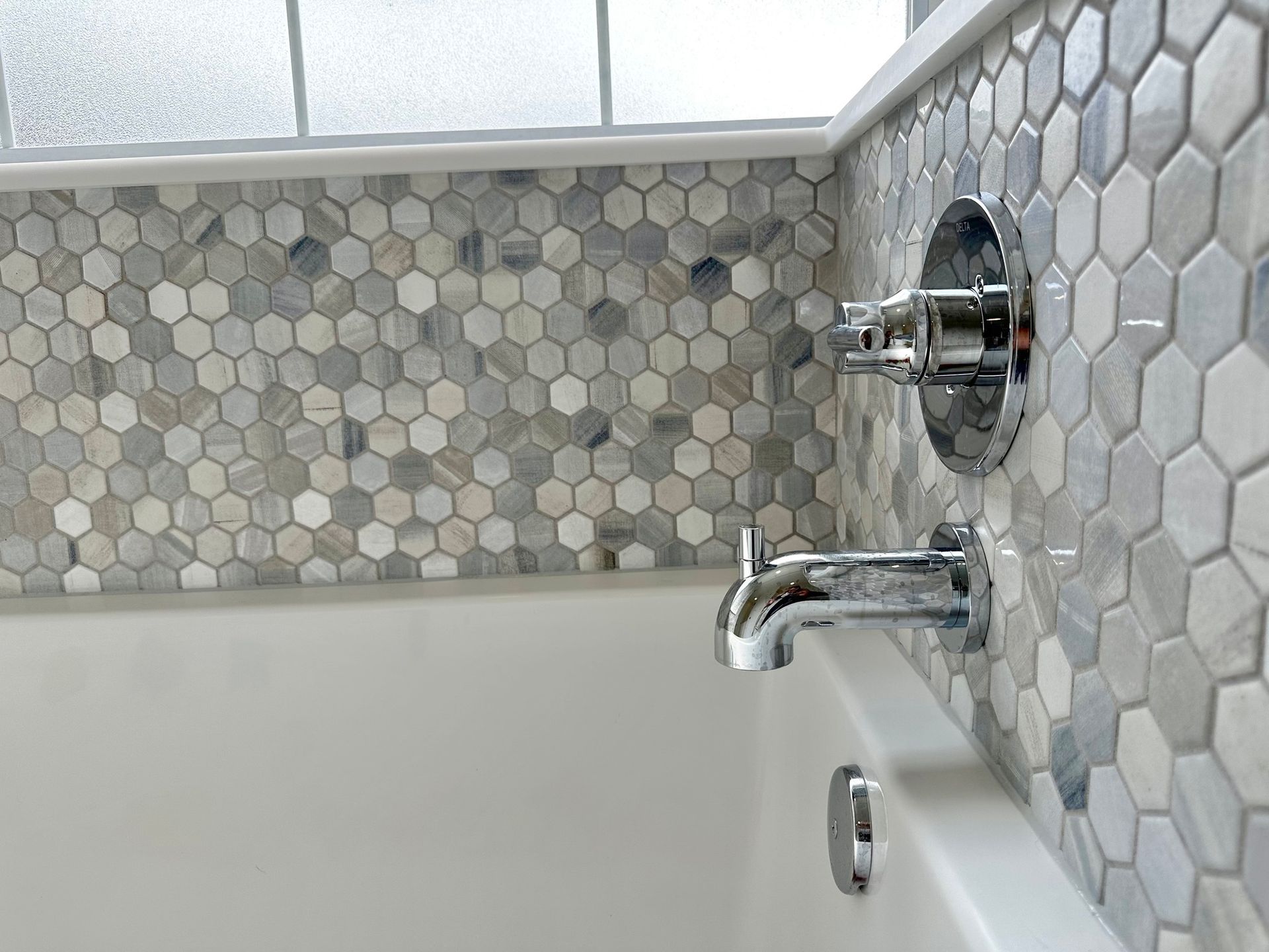 Close-up of a bathtub with mosaic tile wall. Silver fixtures, white tub, and gray, white, and blue hexagonal tiles.