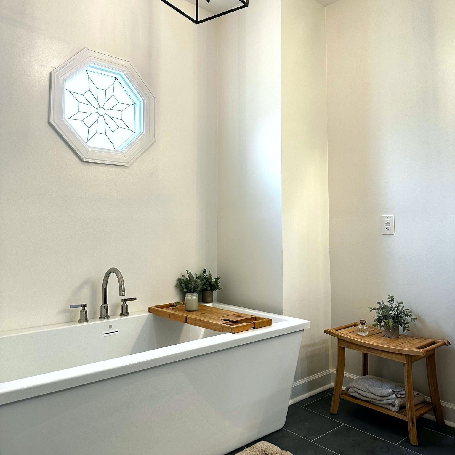 A white clawfoot bathtub in a neutral-toned bathroom with wooden accents, a starburst window, and small plants.