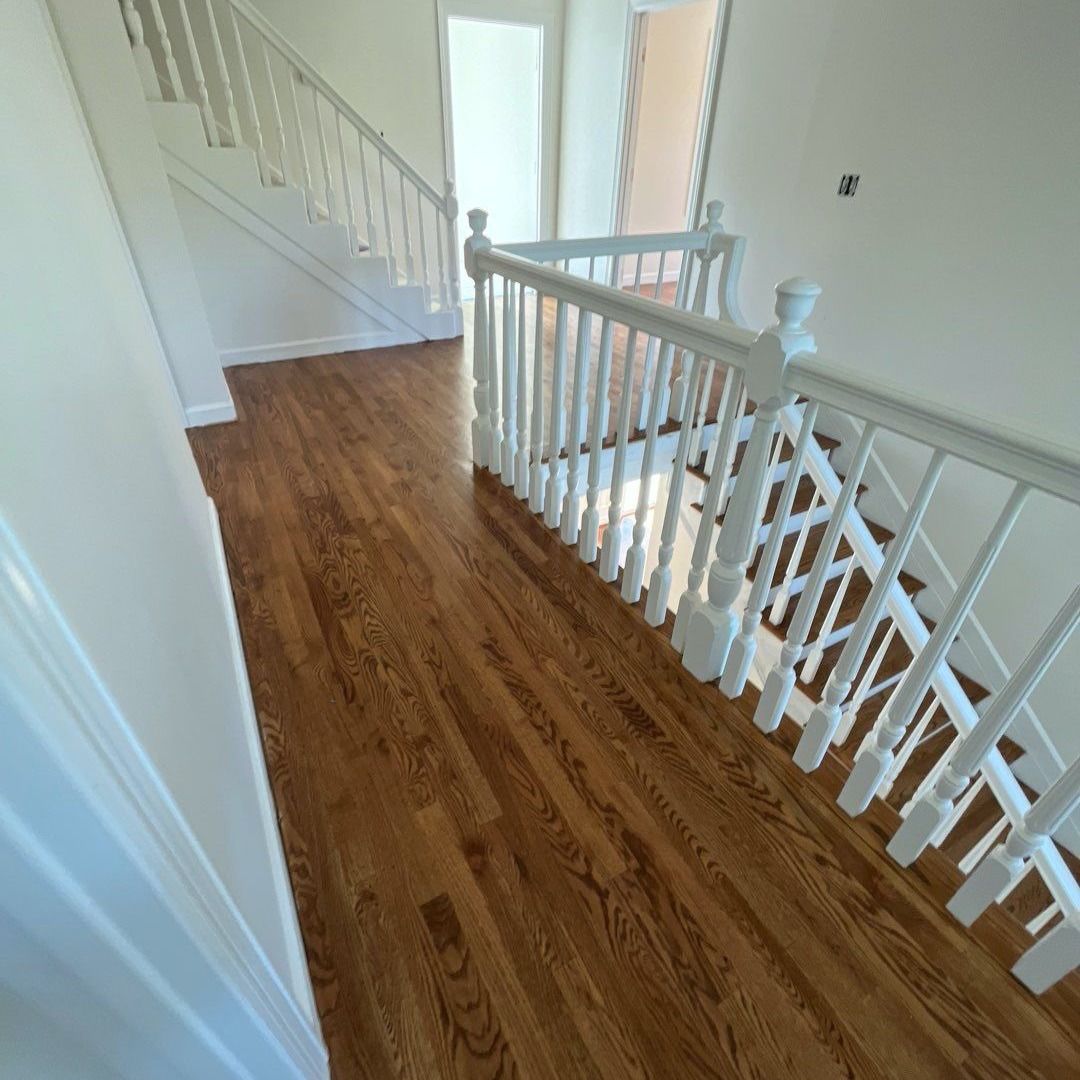 Wooden floor hallway with white staircase railing and a glimpse of another room.