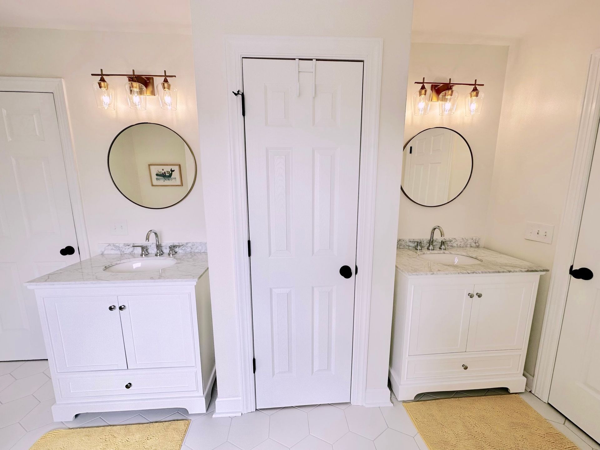 Two white bathroom vanities with round mirrors, sconces, and a door in a bright bathroom.