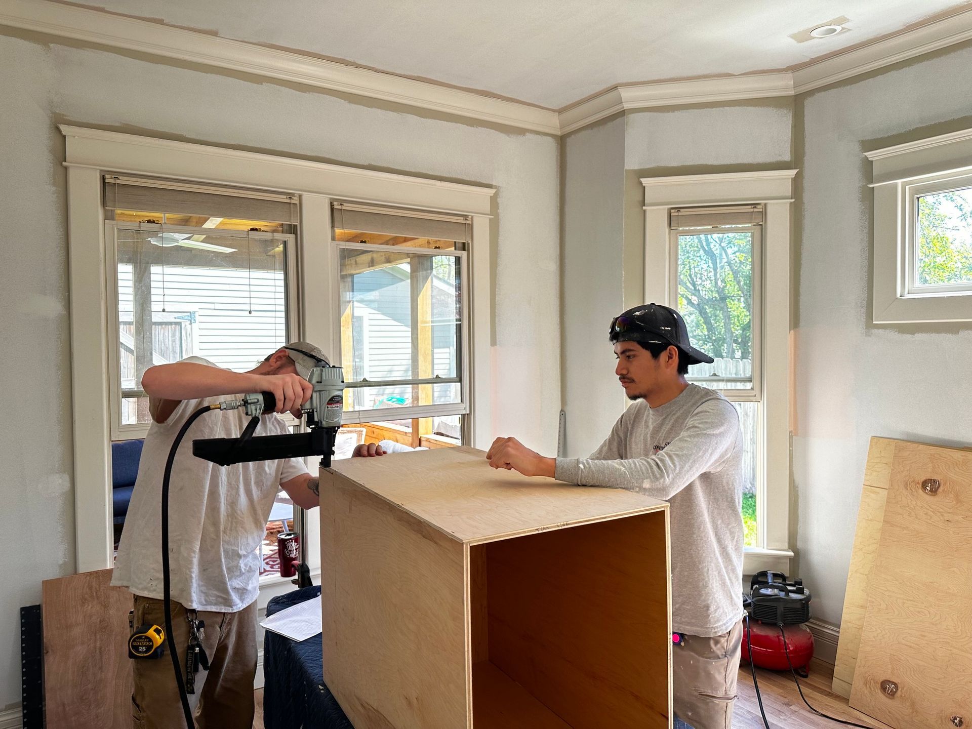 Two workers assembling a wooden cabinet in a room with windows. One uses a nail gun.