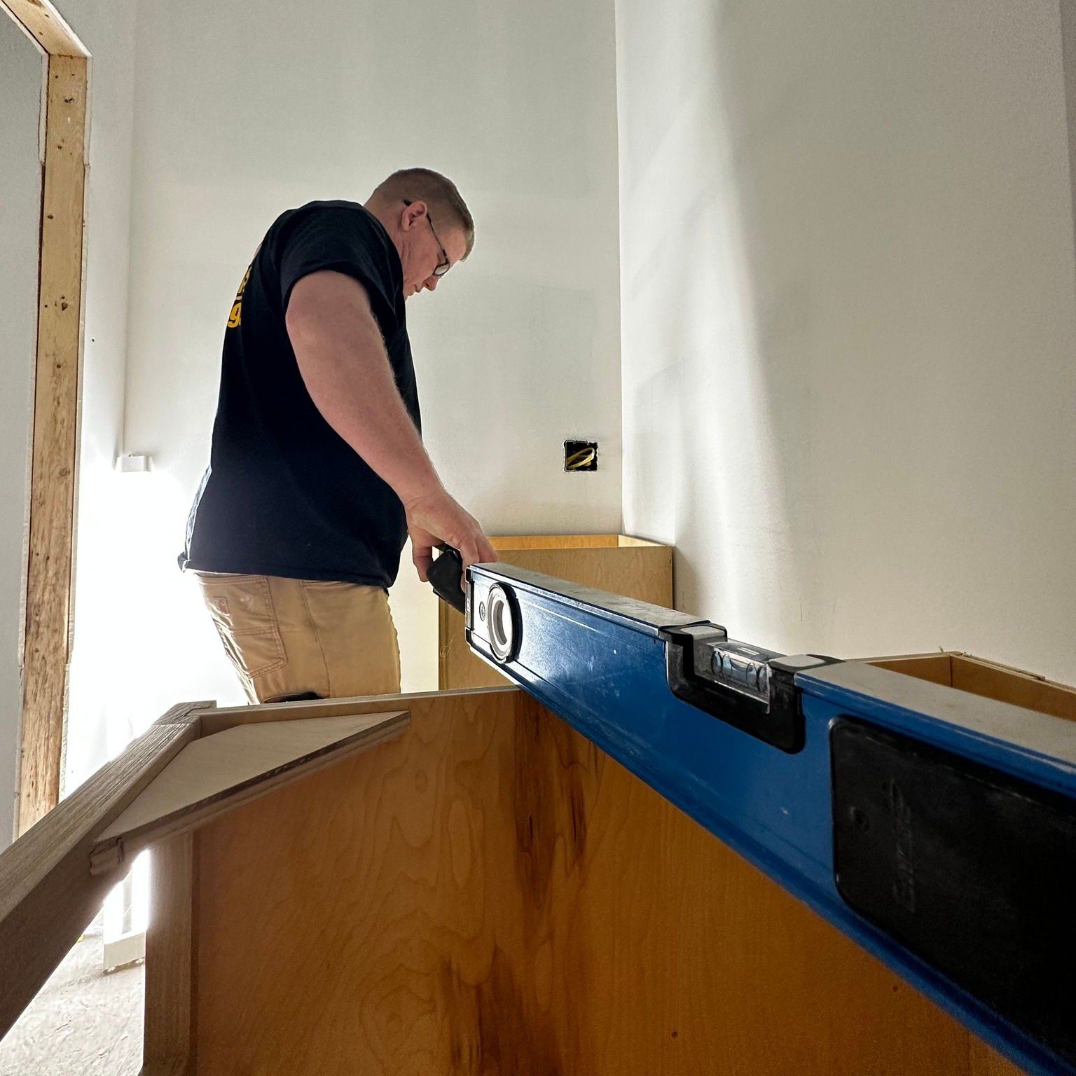 Man in black shirt and khakis using a level on a wooden structure, possibly cabinet installation. White wall.